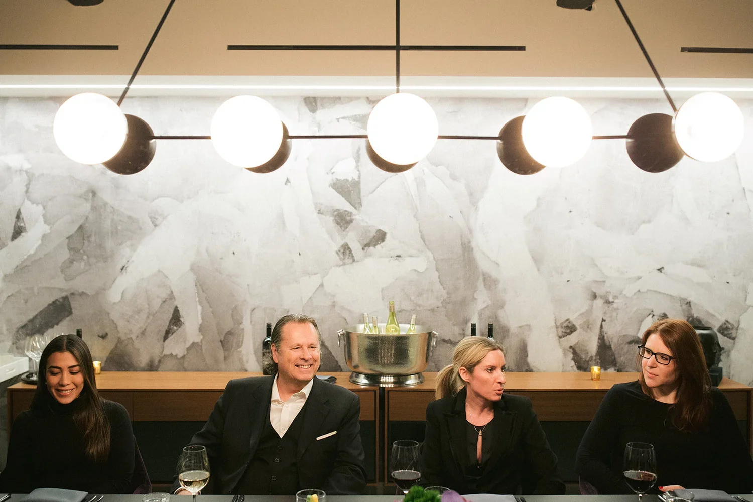 Four people seated at a dinner table with glasses of wine, indoors with modern lighting and a textured wall background.
