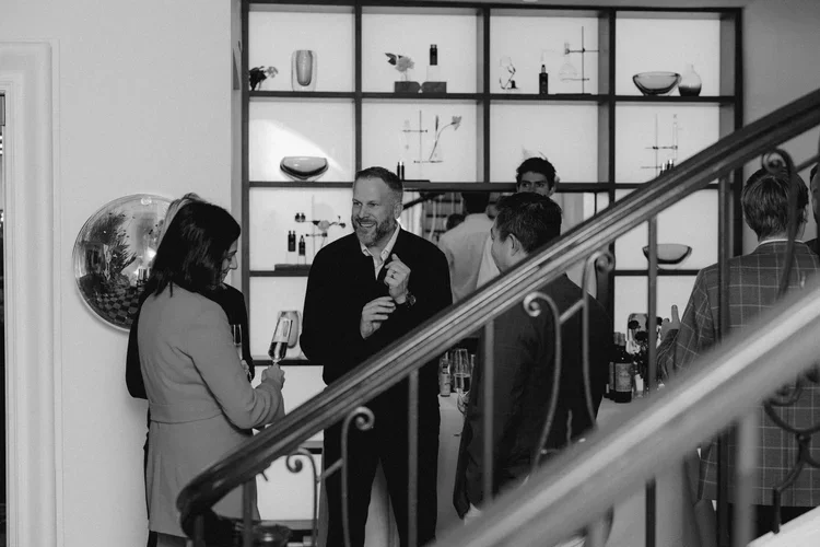 People socializing at a gathering or party, with some holding drinks, in a modern indoor setting with shelves displaying decorative items and art.
