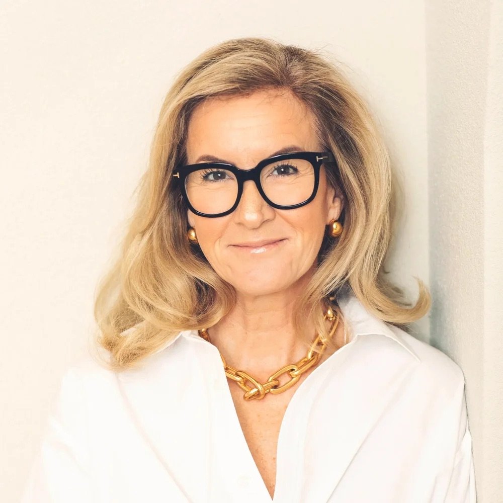 A woman with blonde hair, wearing black glasses, gold earrings, a gold chain necklace, and a white blouse, smiling and leaning against a white wall.