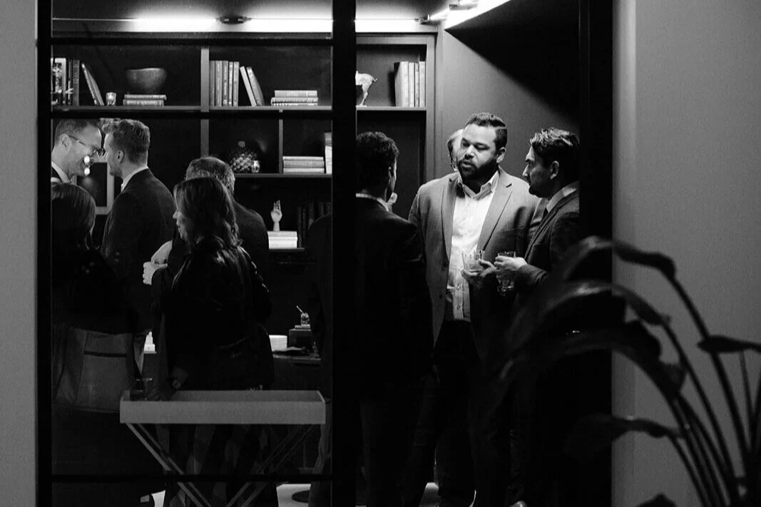 People socializing in a modern office or lounge, seen through a glass window, with a bookshelf and a plant in the background.