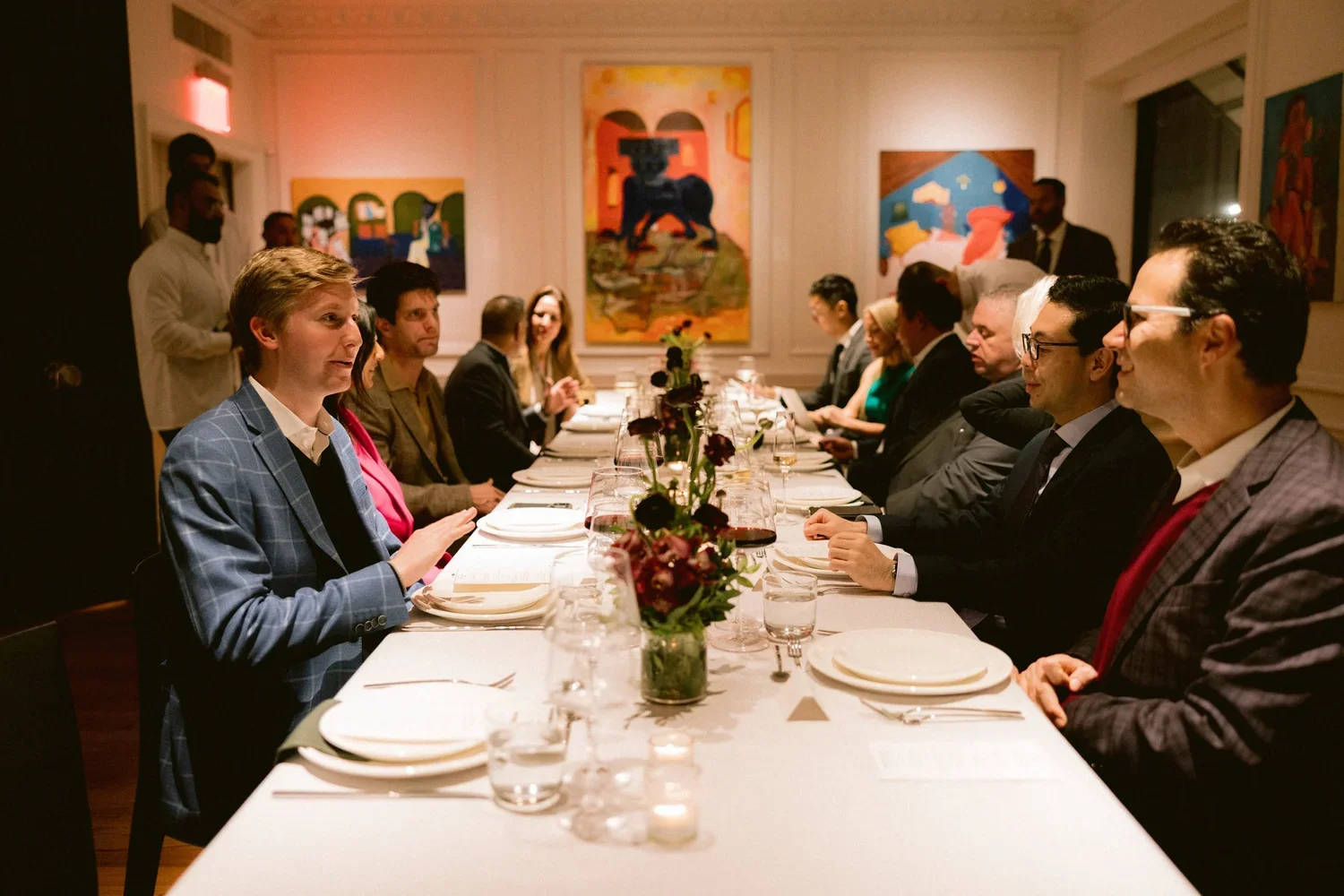 Business people seated at a long dining table in a restaurant or private dining room with colorful artwork on the walls.
