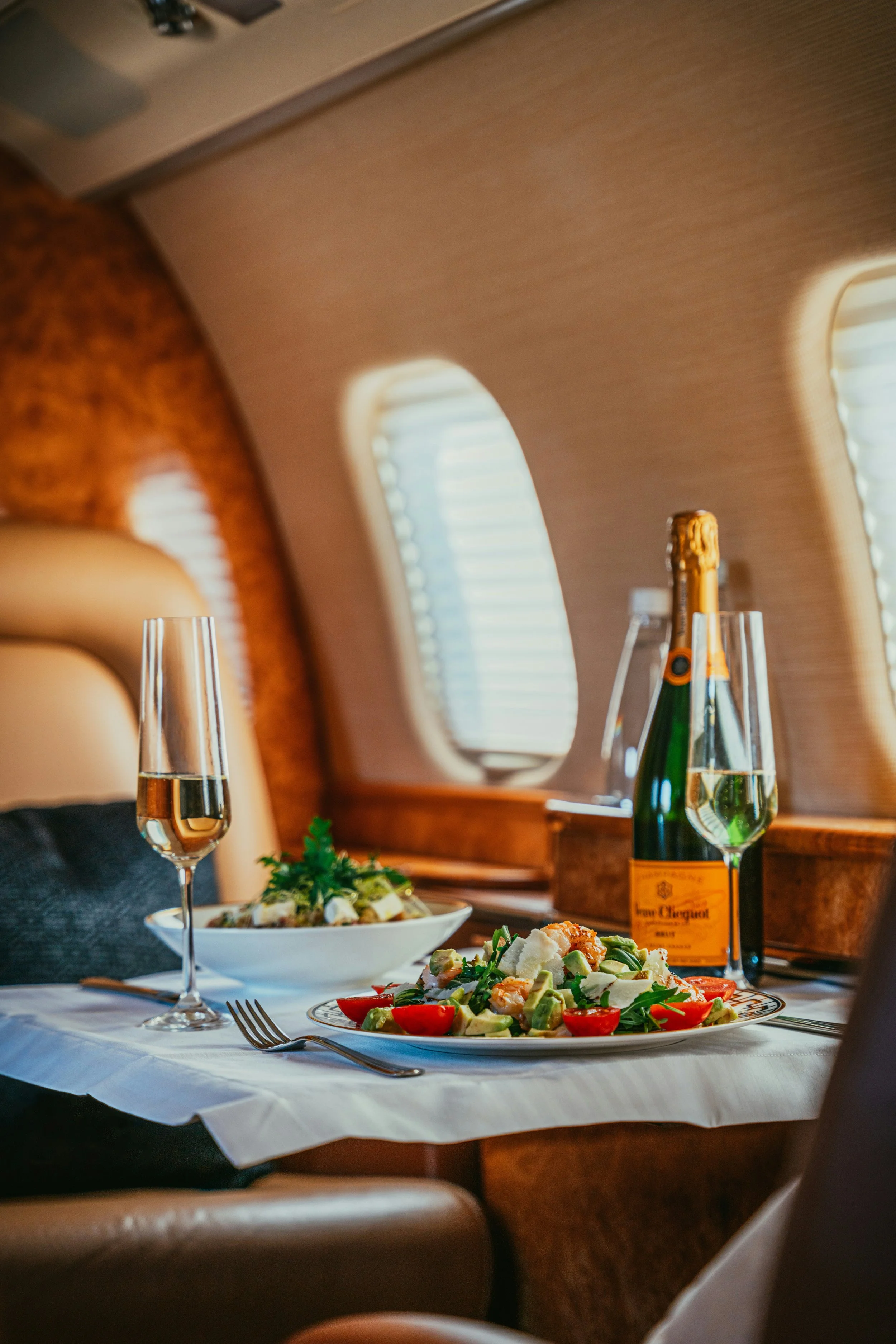 A dining scene inside a private jet with a table featuring a bottle of champagne, two glasses, and plates of salad and seafood. The interior has beige walls and small rectangular windows with blinds.