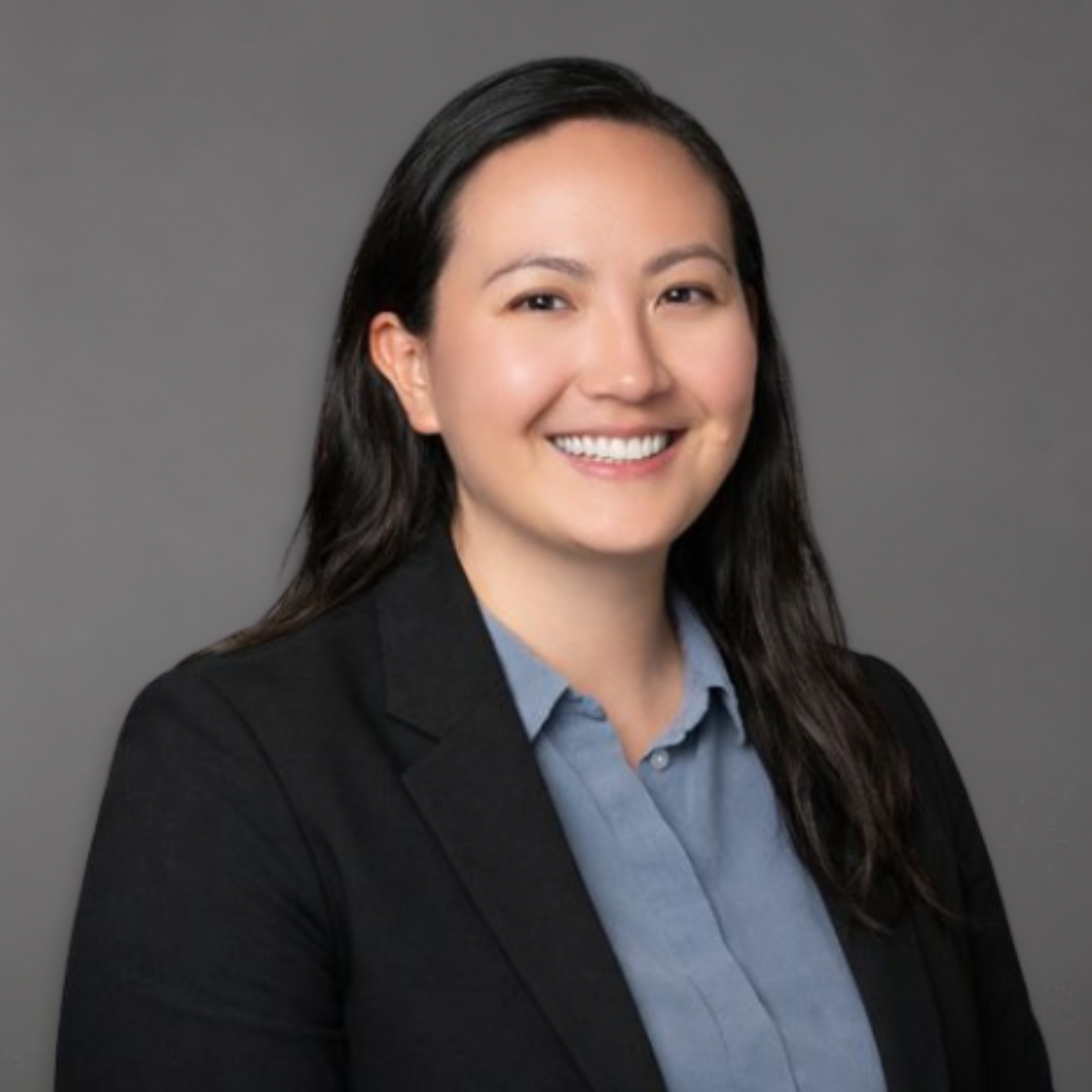 A woman with long dark hair smiling, wearing a black blazer and a blue collared shirt, against a plain gray background.