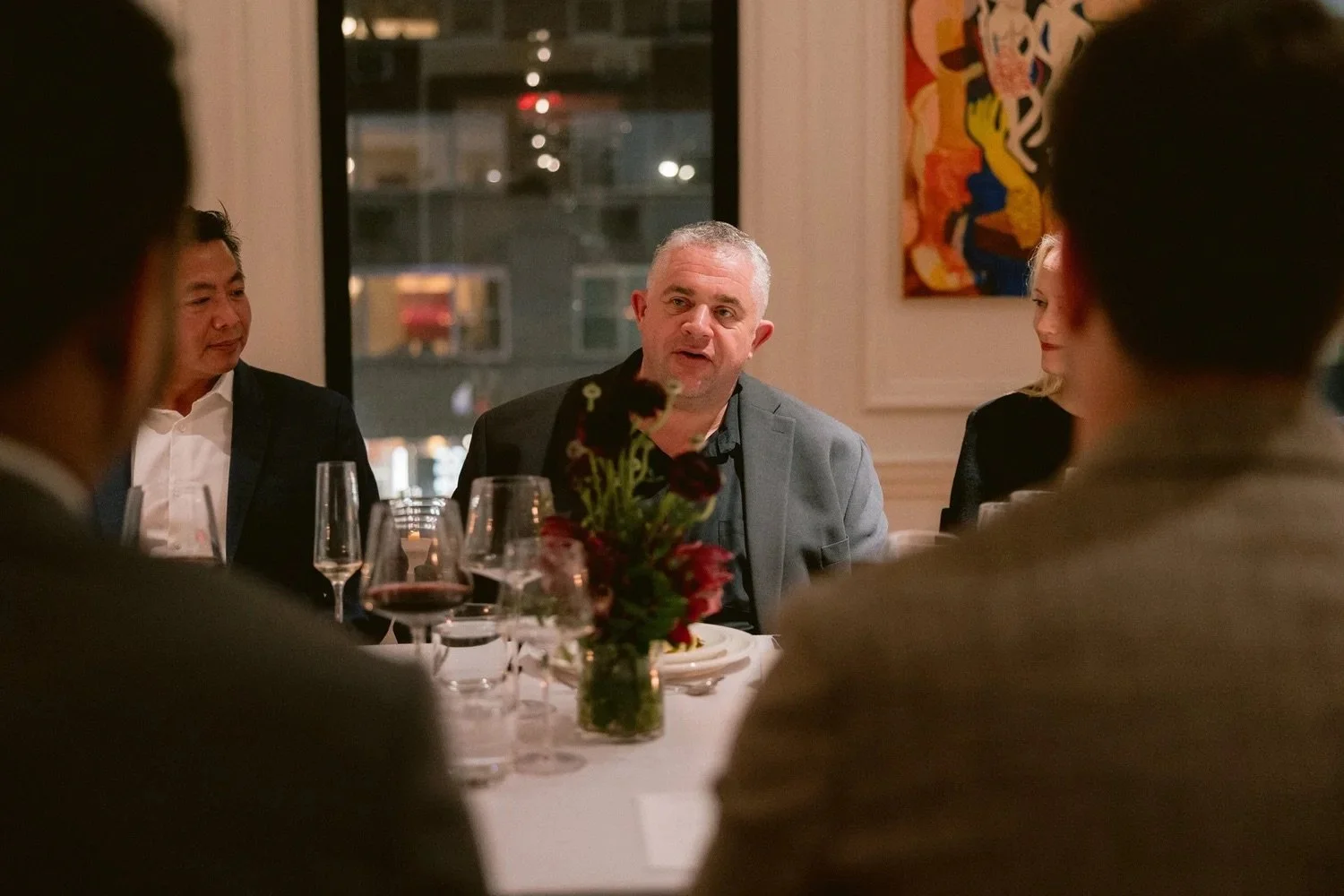 A man in a grey blazer speaking at a dinner table, with five people seated around, glasses of red wine, a floral centerpiece, and a colorful abstract painting on the wall.