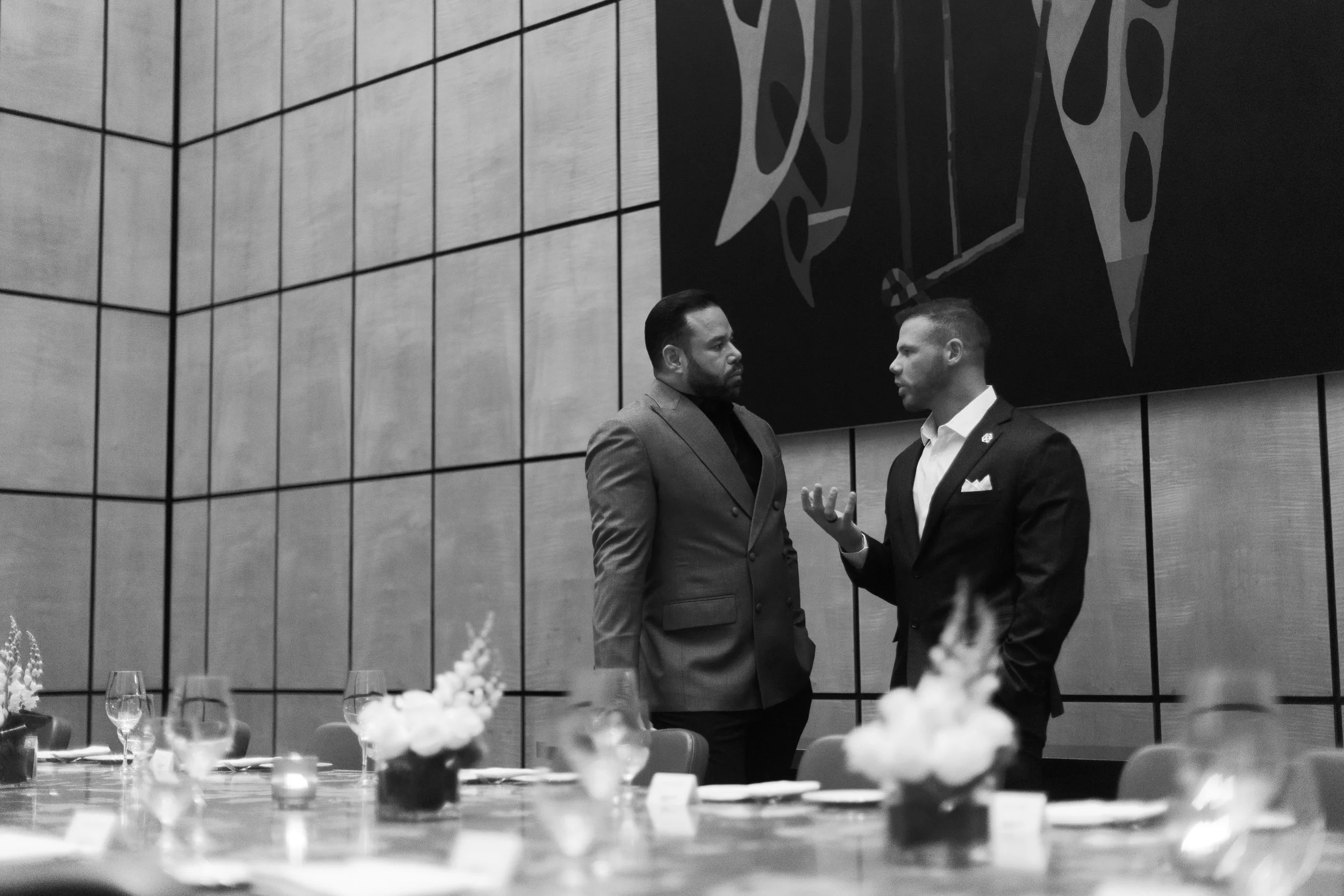 Two men in suits are engaged in a conversation at a formal event, standing in front of a large wall with artwork. The foreground shows a table with glassware and floral arrangements.