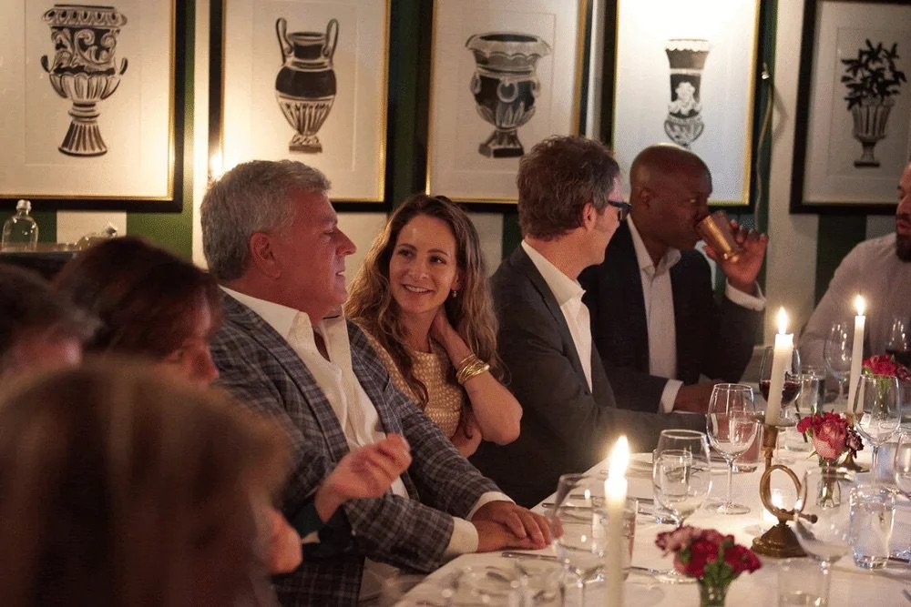 People sitting at a dinner table with lit candles and wine, engaged in conversation, in a warmly lit room with framed artwork of vases on the wall.