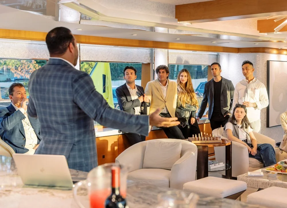 A man in a checkered blazer giving a presentation to a group of young professionals in a modern, well-lit office or conference room with large windows.