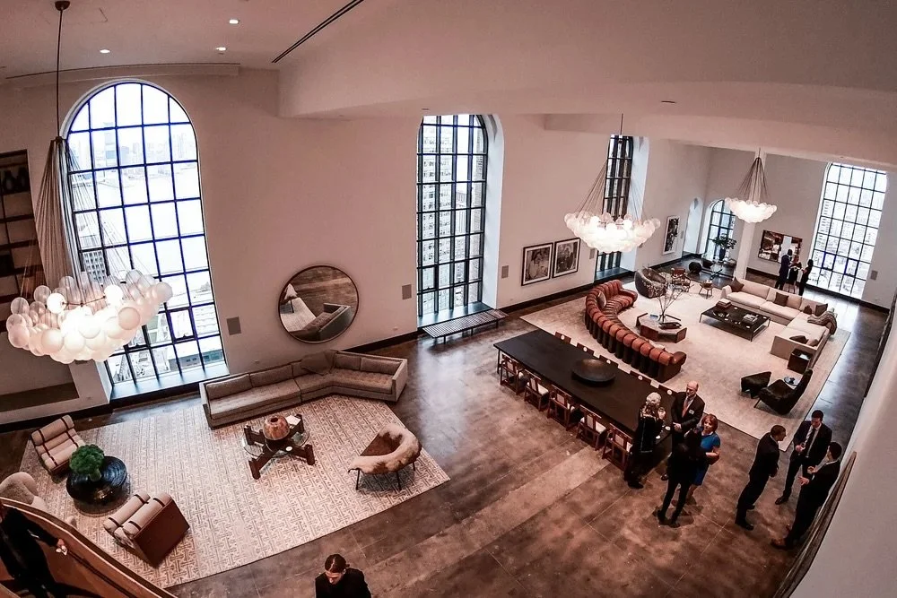 Large modern lounge with high ceilings, large arched windows, and contemporary furniture. Several groups of people are socializing near the bar and seating areas.