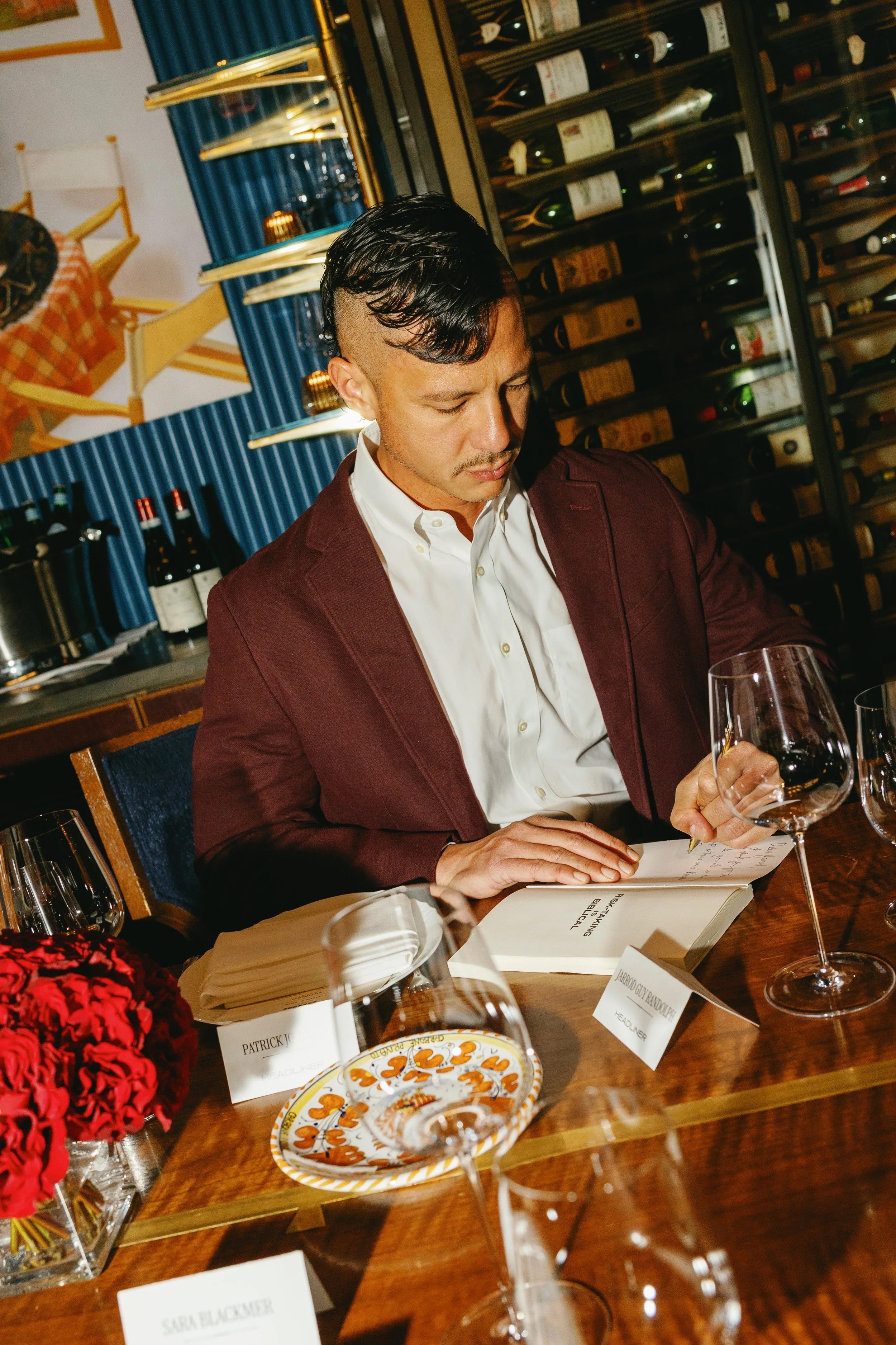 A man dressed in a maroon blazer and white shirt sitting at a table in a restaurant or wine cellar, reading a document. The table has glasses of wine, a plate with cookies, and a red flower centerpiece. There is a wine rack filled with bottles behind him.