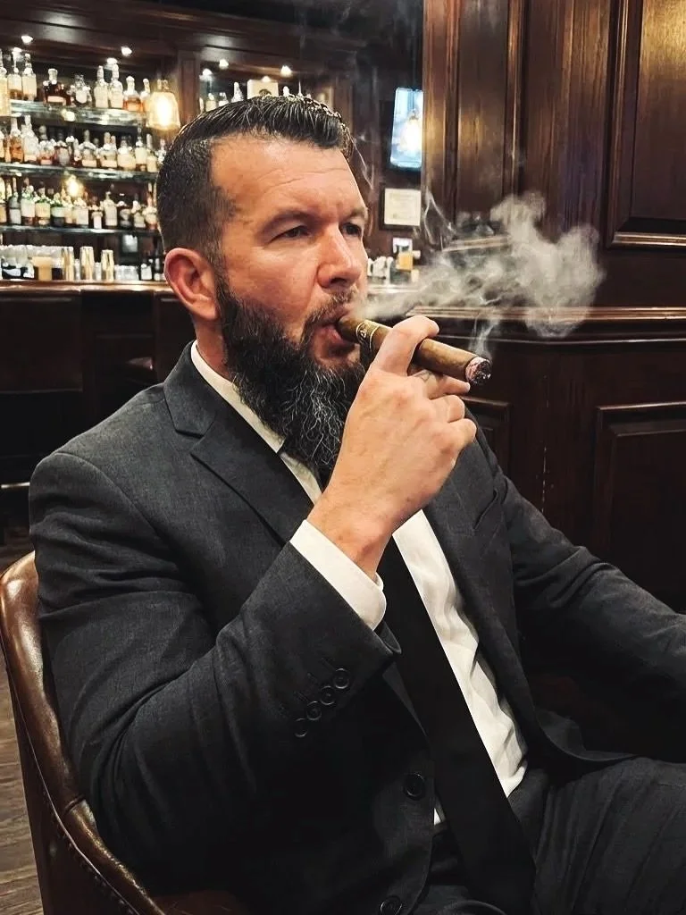 A man dressed in a suit with a beard and slicked-back hair is sitting in a bar, smoking a cigar, with a background of shelves filled with bottles.