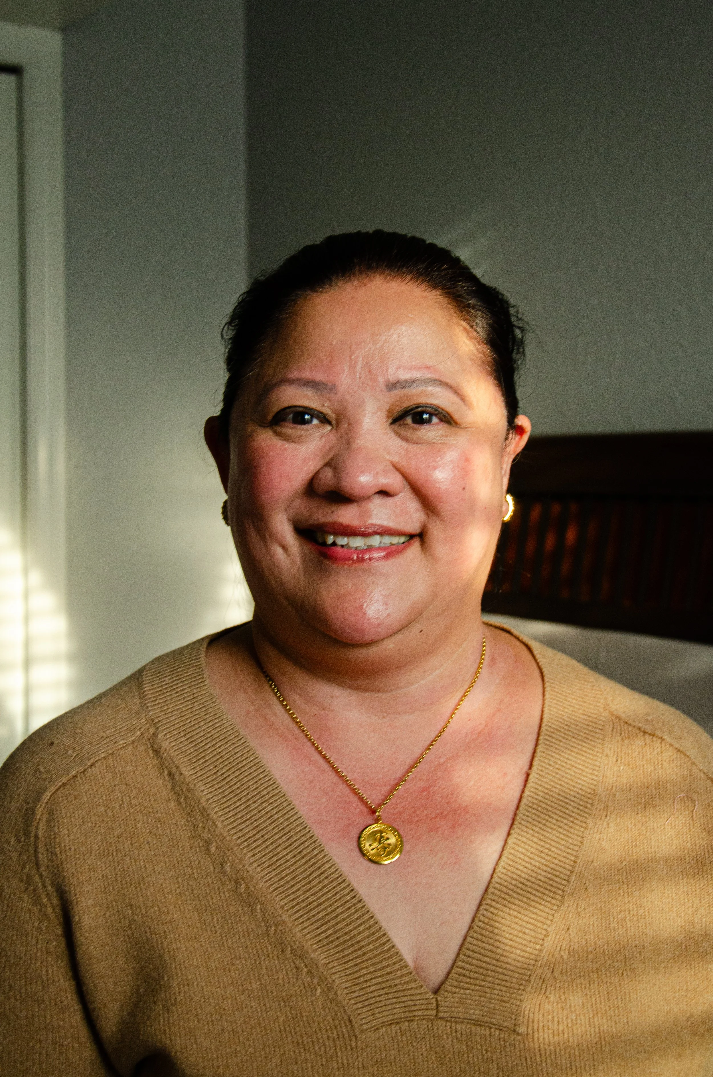 Close-up of a smiling woman with dark hair pulled back, wearing a beige V-neck sweater and gold jewelry, standing indoors with gray walls and a window with blinds in the background.