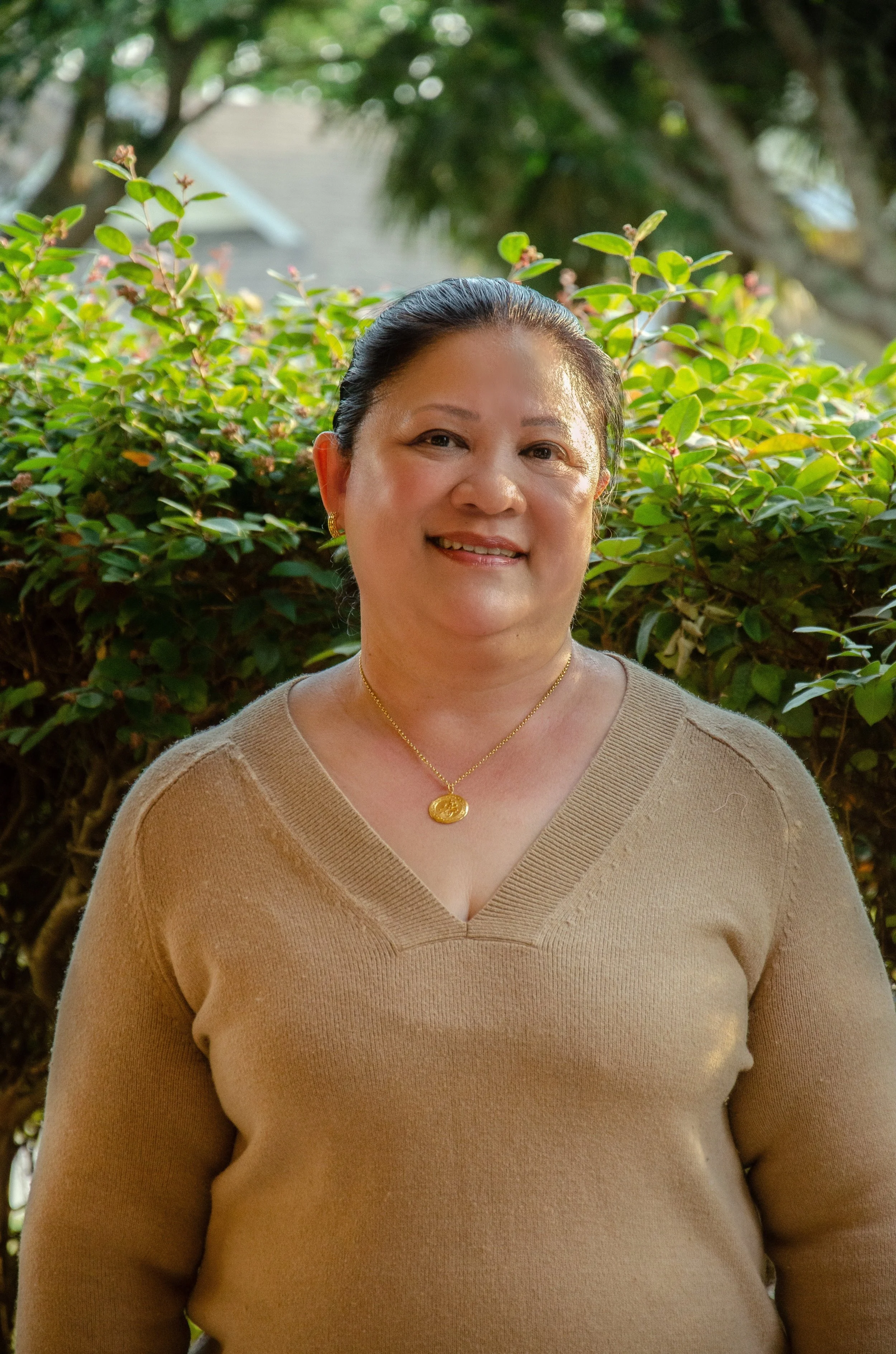 A woman with dark hair tied back, wearing a beige V-neck sweater, gold earrings, and a gold necklace, smiling outdoors with greenery in the background.