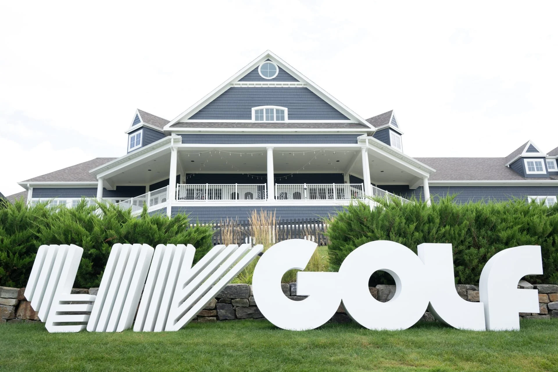 Large white sign spelling 'WELL GOLF' in front of a modern blue house with a porch, green bushes, and a stone wall, under a cloudy sky.