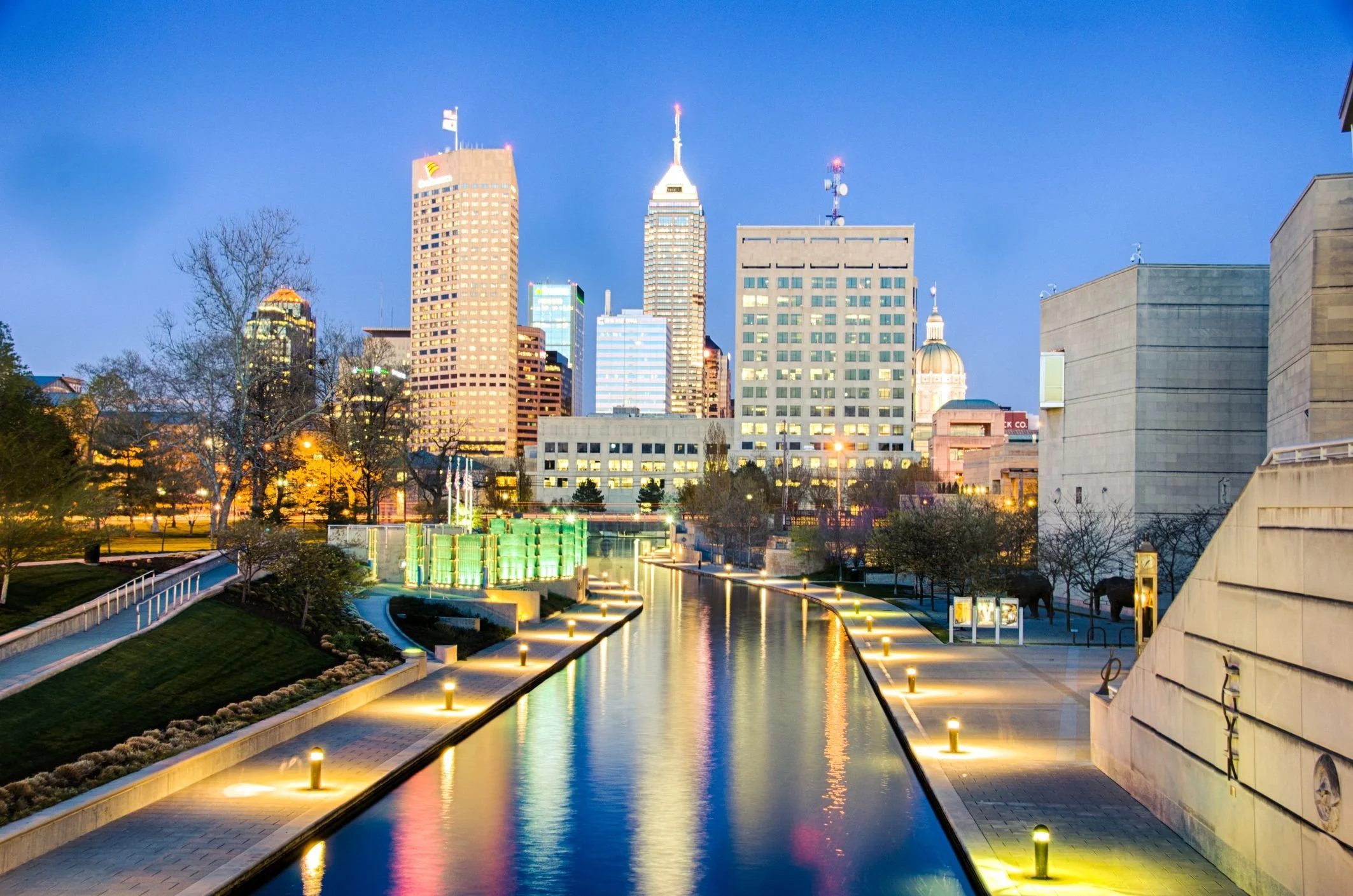 City skyline at dusk with illuminated buildings, a canal with reflections, and lit pathways, in Indianapolis.