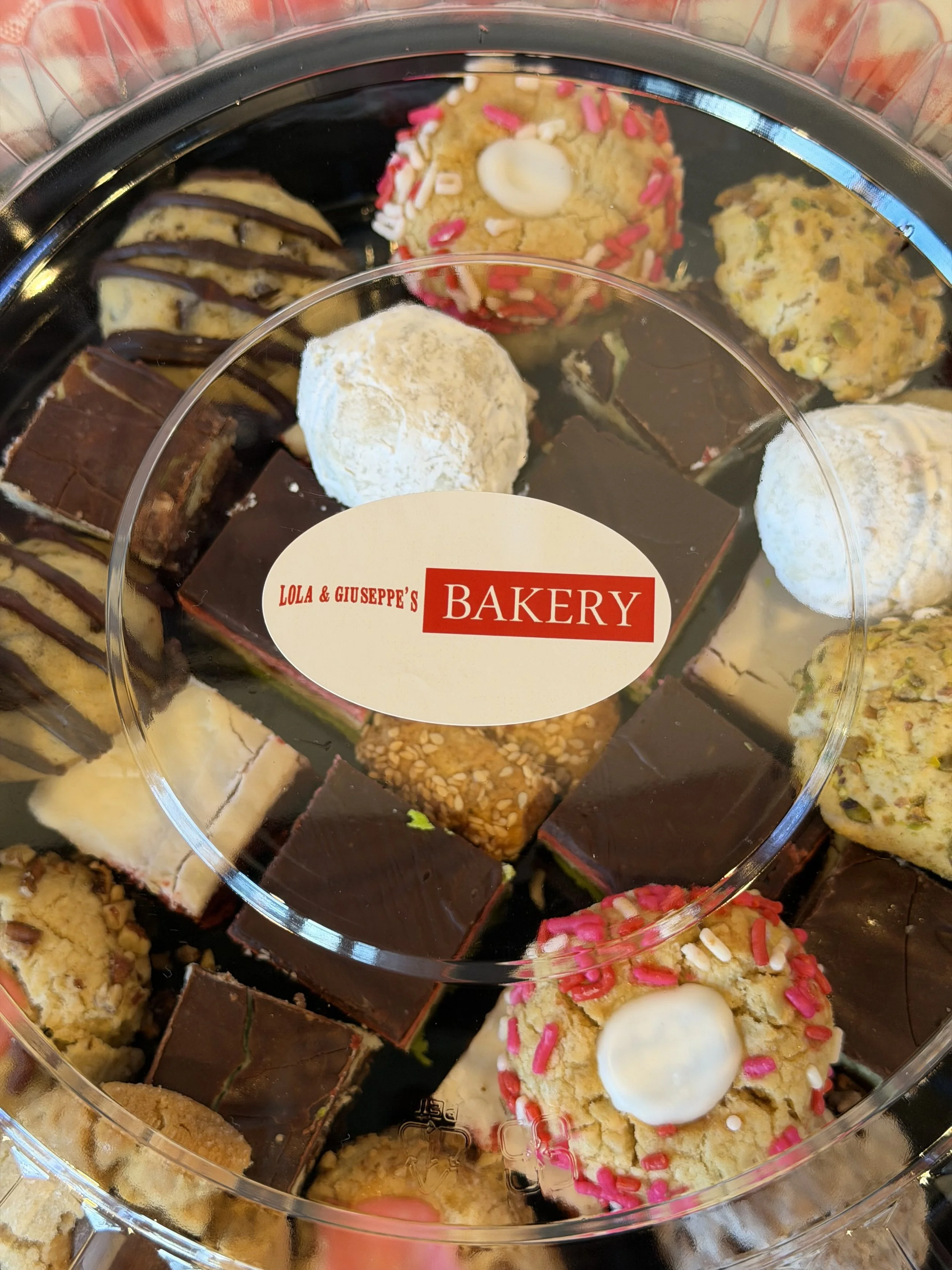 An assortment of various cookies in a clear plastic container, including frosted and sprinkled cookies, chocolate-covered cookies, and cookies with chopped nuts.