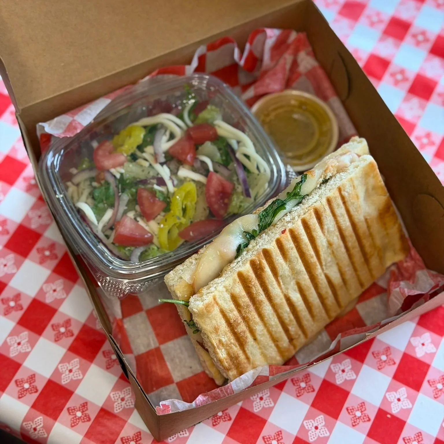 A takeout box with a side salad in a clear plastic container, a grilled panini sandwich with melted cheese and vegetables, and a small cup of dipping sauce, all on red and white checkered paper.