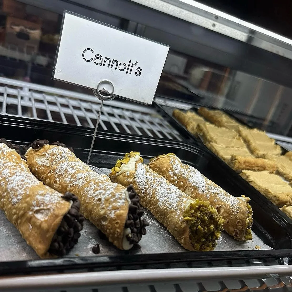 Cannoli in a display case, with some topped with chocolate chips and others with pistachios, dusted with powdered sugar.