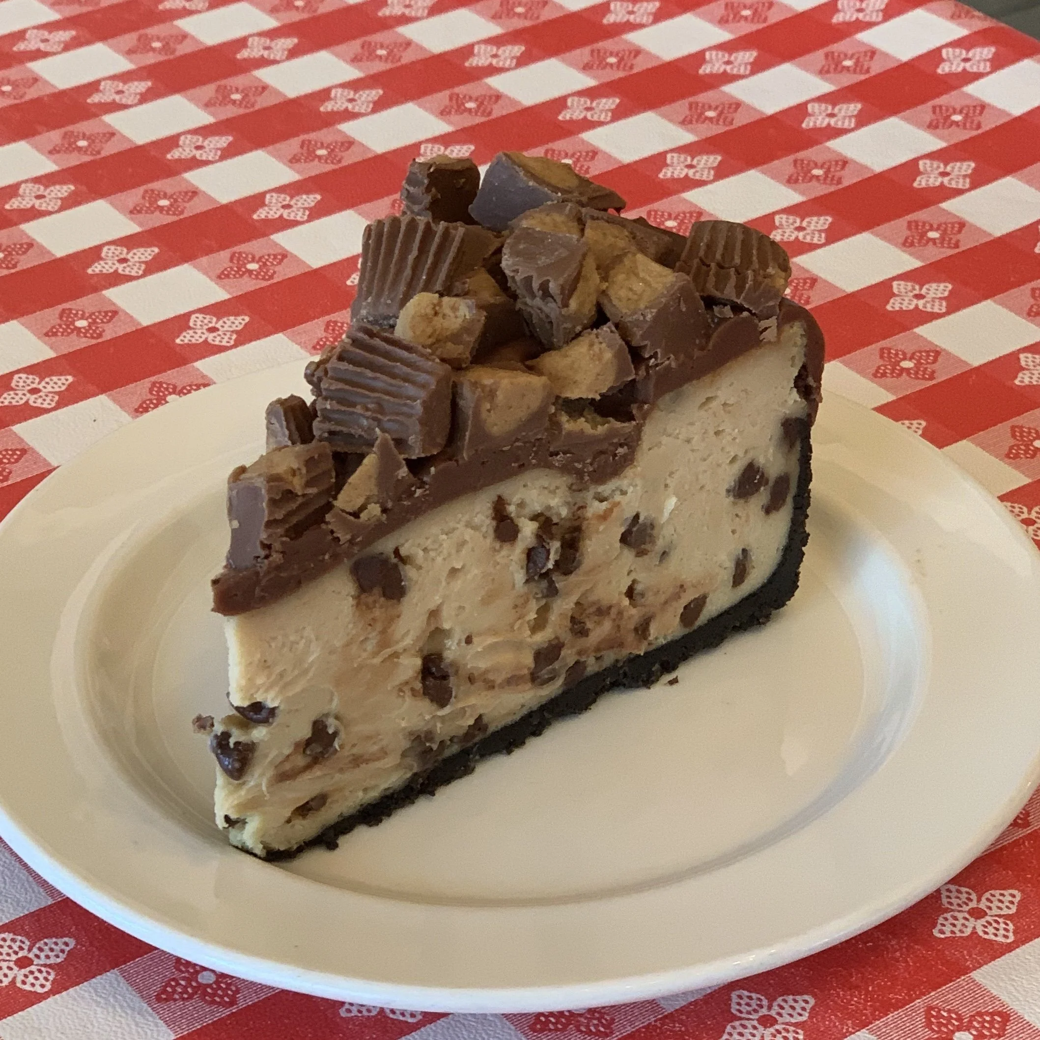 A slice of cheesecake topped with pieces of chocolate and nuts, placed on a white plate on a red and white checkered tablecloth.