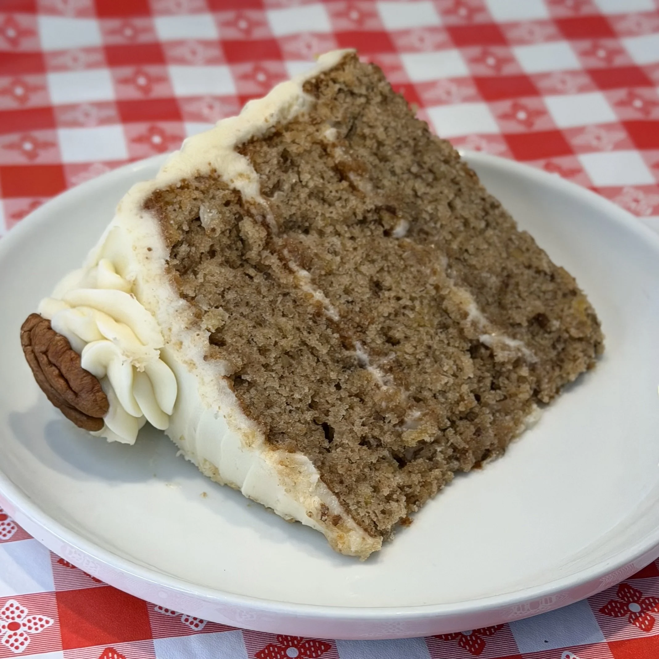 A slice of pecan and possibly banana bread with a thick layer of vanilla frosting, served on a white plate, with a red and white checkered tablecloth in the background.