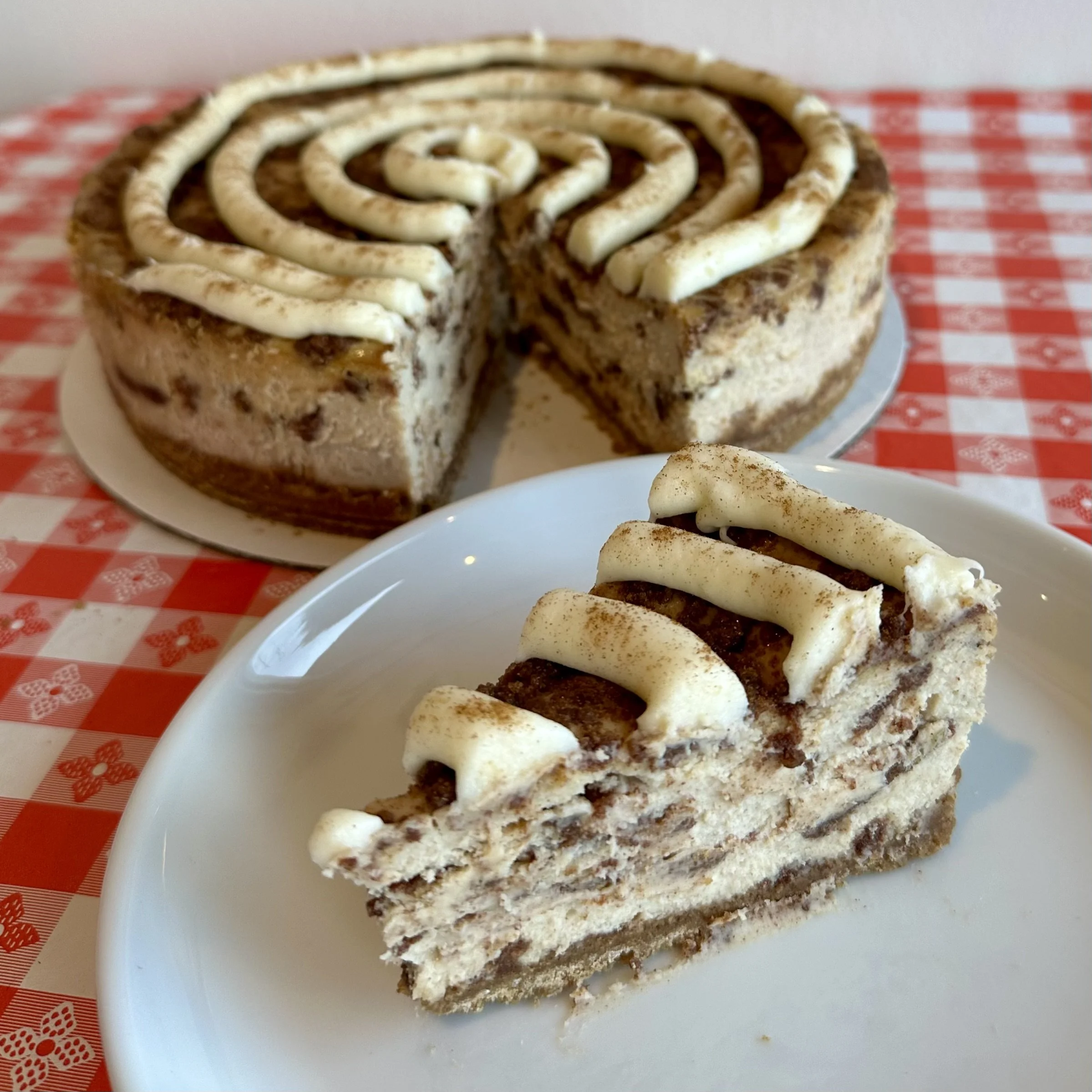 Close-up of a slice of cinnamon swirl coffee cake on a white plate, with a fork partially visible in the foreground, placed on a checkered red and white tablecloth.