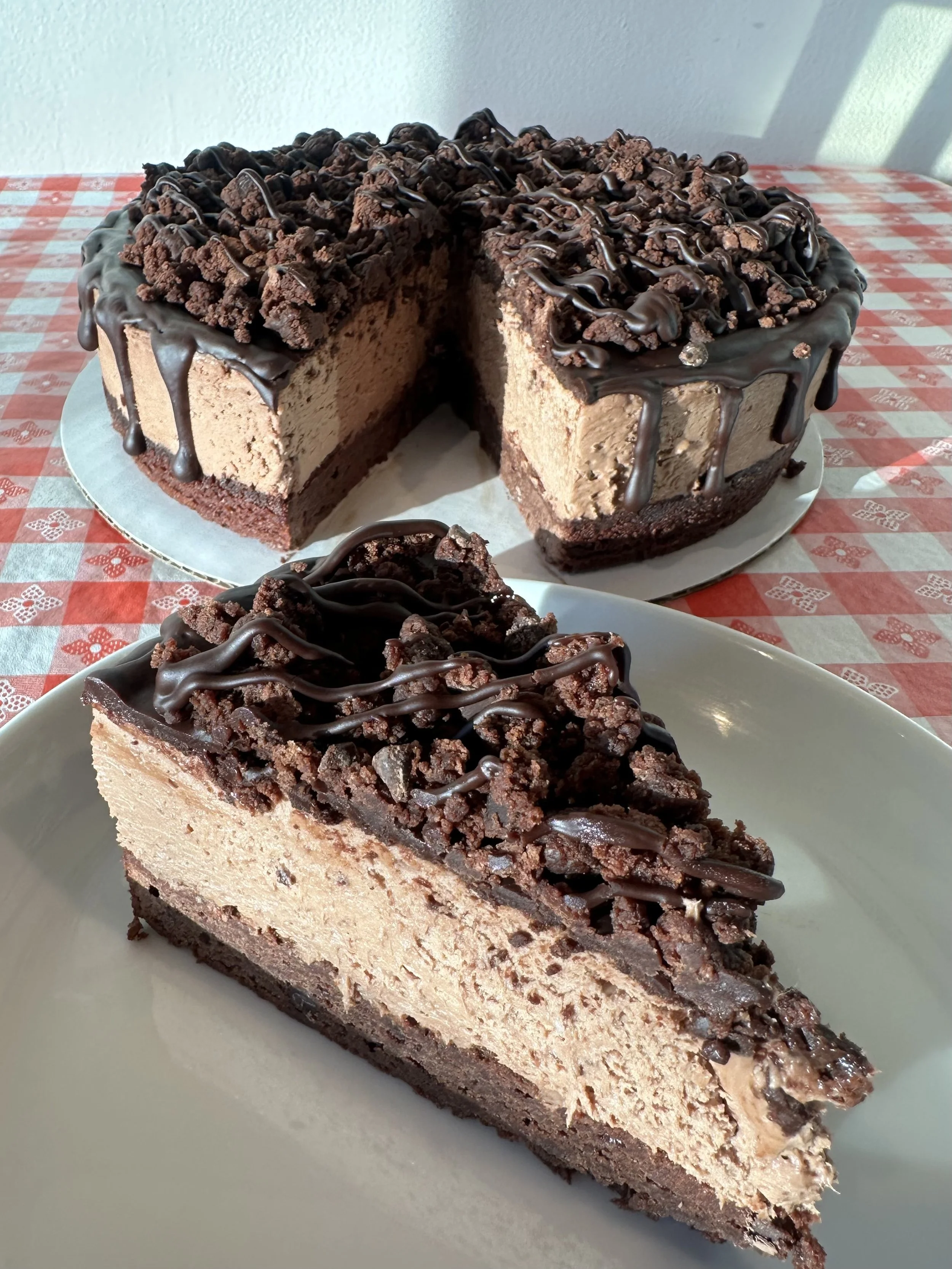 A slice of chocolate mousse cake with a chocolate crust, layered with whipped chocolate mousse, topped with crumbled chocolate pieces and a drizzle of chocolate syrup, on a light-colored plate.