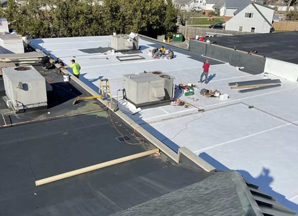 Workers installing a white reflective roofing material on a building's roof, with various tools and equipment scattered around.