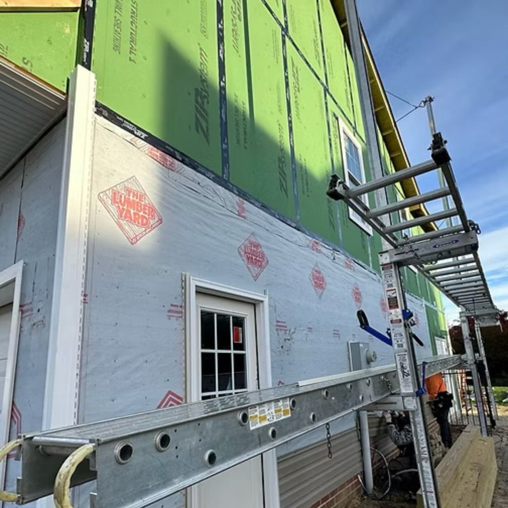 Building under construction with green sheathing on upper level, white sheathing on lower level, and a metal ladder leaning against the wall. There is a worker in an orange shirt near the base of the ladder.