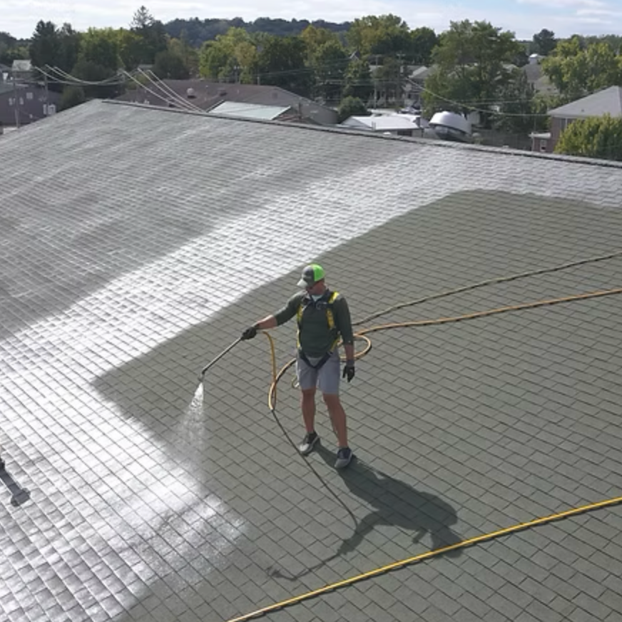 Person cleaning a large rooftop with a hose, wearing gloves, a cap, and a harness, in a residential neighborhood with trees and houses in the background.