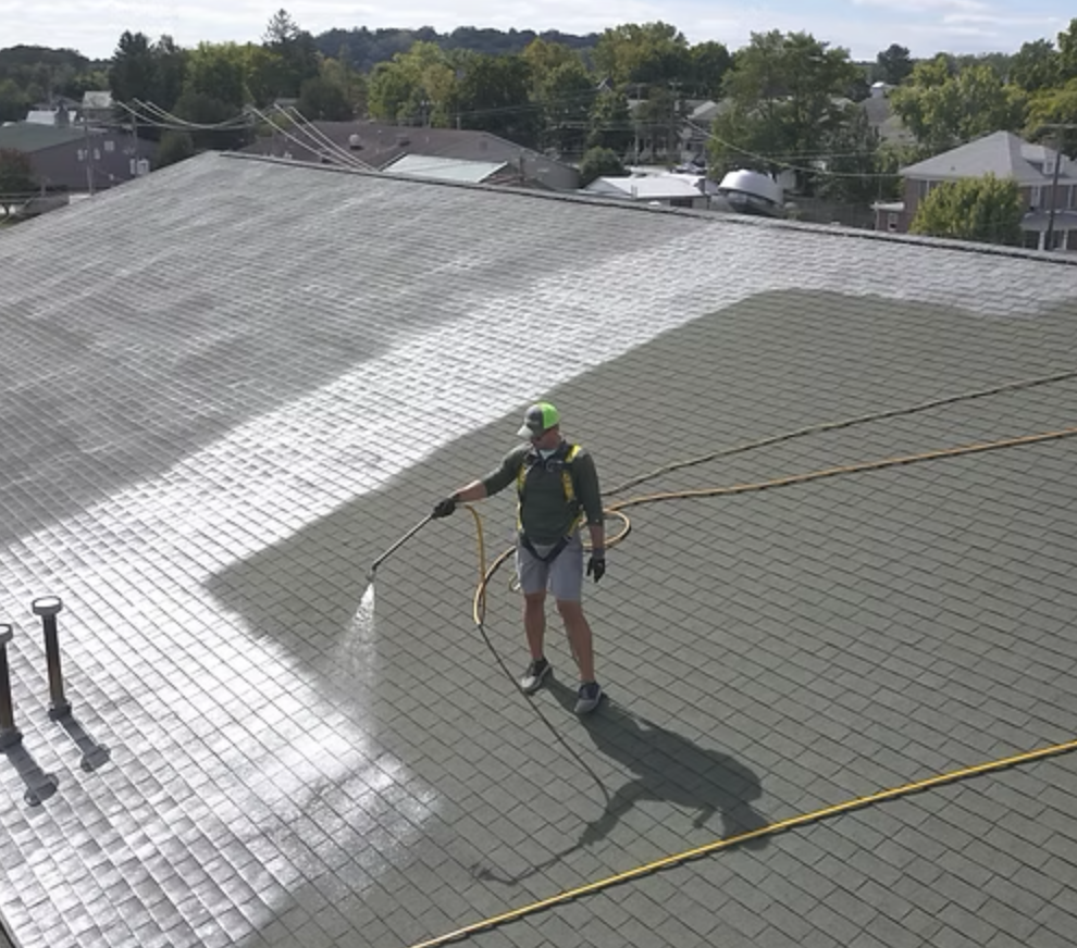 Person wearing a green cap and spraying a roof with a hose, with houses and trees in the background.