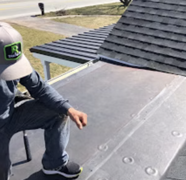 Person sitting on a bench on a rooftop, wearing a cap and jeans, with a house roof and driveway visible in the background.