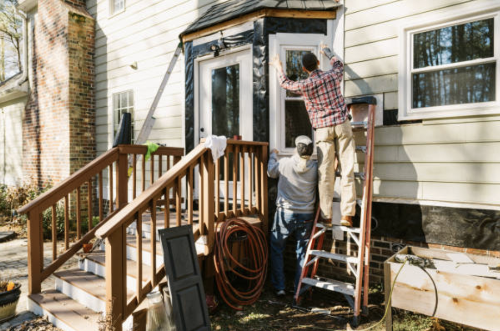 Two people working on home renovation, painting and installing siding on the front door area of a house with beige siding, wooden stairs, and a railing.
