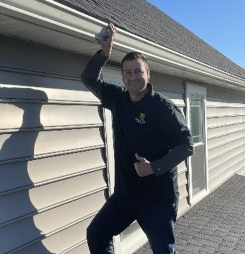 A man standing outside on a deck in front of a house, smiling, holding a cup up near the roof, giving a thumbs-up with the other hand, during daytime with blue sky.