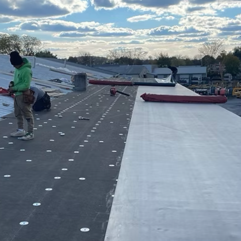Roof workers installing a new roofing material on a sloped roof during daytime, with a partly cloudy sky overhead.