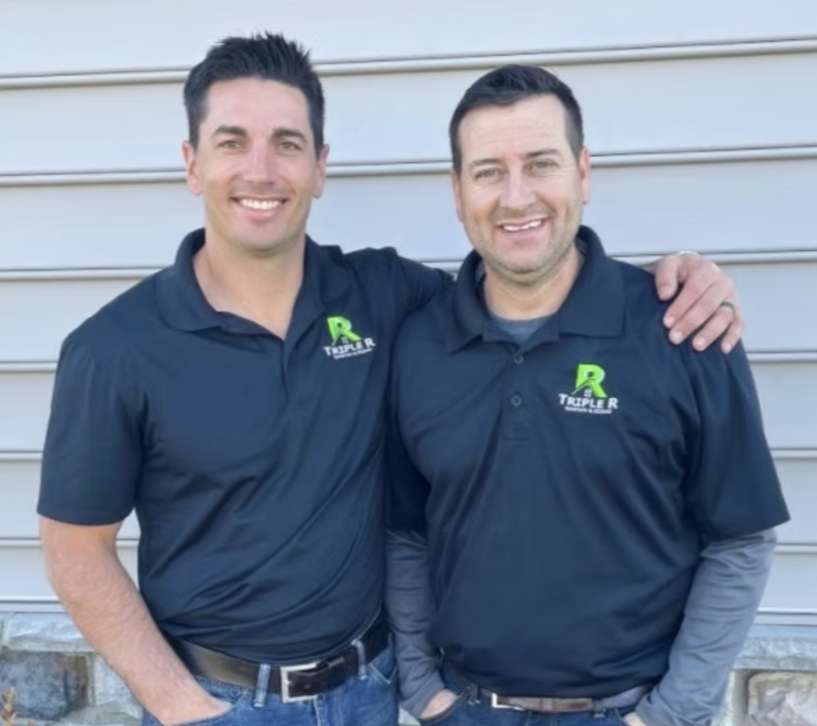 Two men standing side by side, smiling, wearing matching dark blue polo shirts with a green and white logo that says 'Triple R'. One has his arm around the other's shoulder, and they are standing in front of a grey, horizontal metal siding wall.