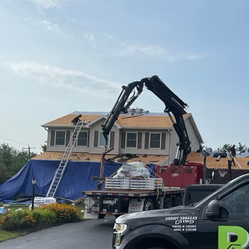 Construction workers work on the roof of a house as a crane lifts roofing materials. The house is partially covered with blue tarp and surrounded by some flowers.