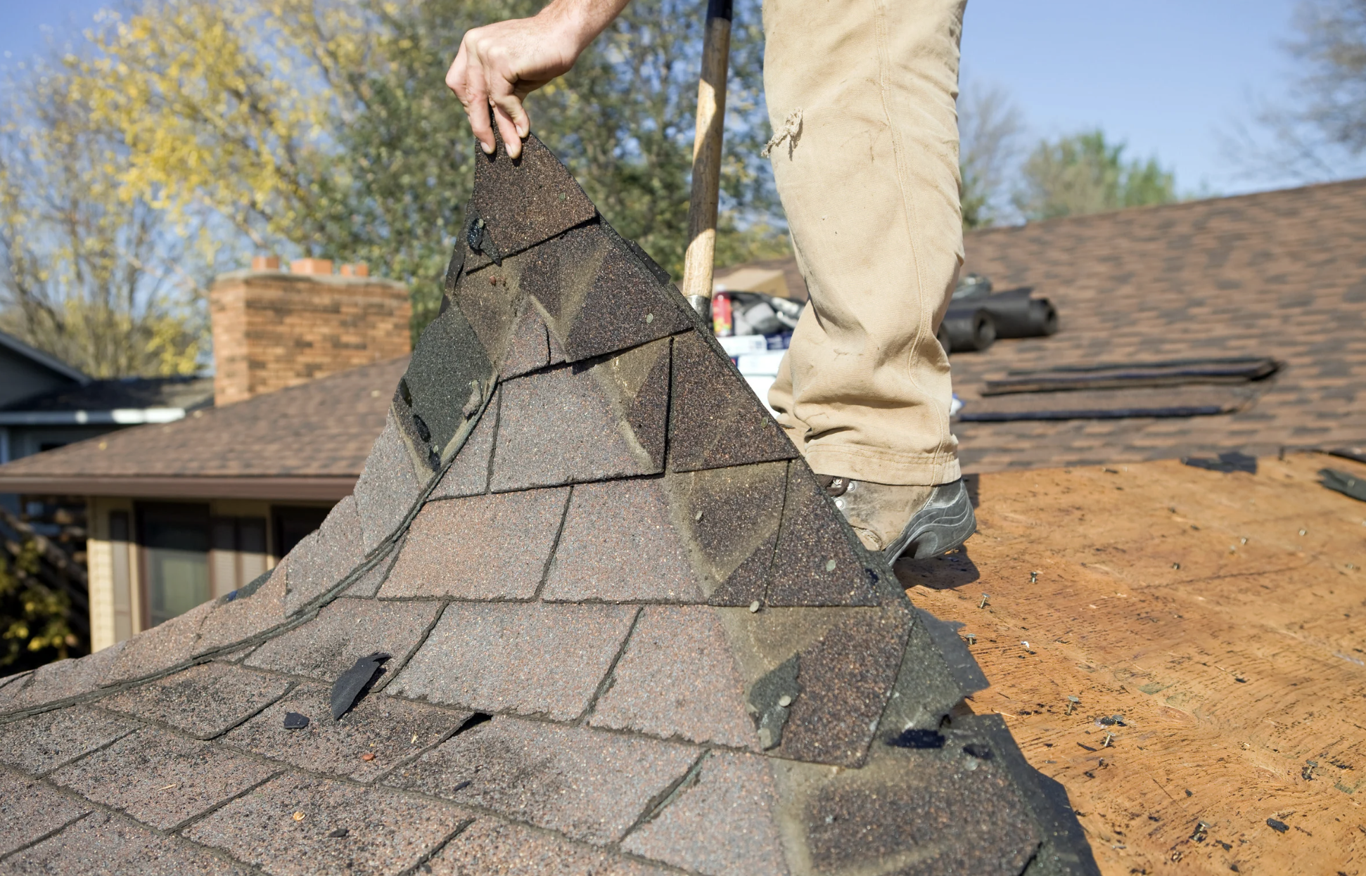 A person working on a roof, lifting and adjusting asphalt shingles during roof repair or replacement with a background of house, trees, and blue sky.