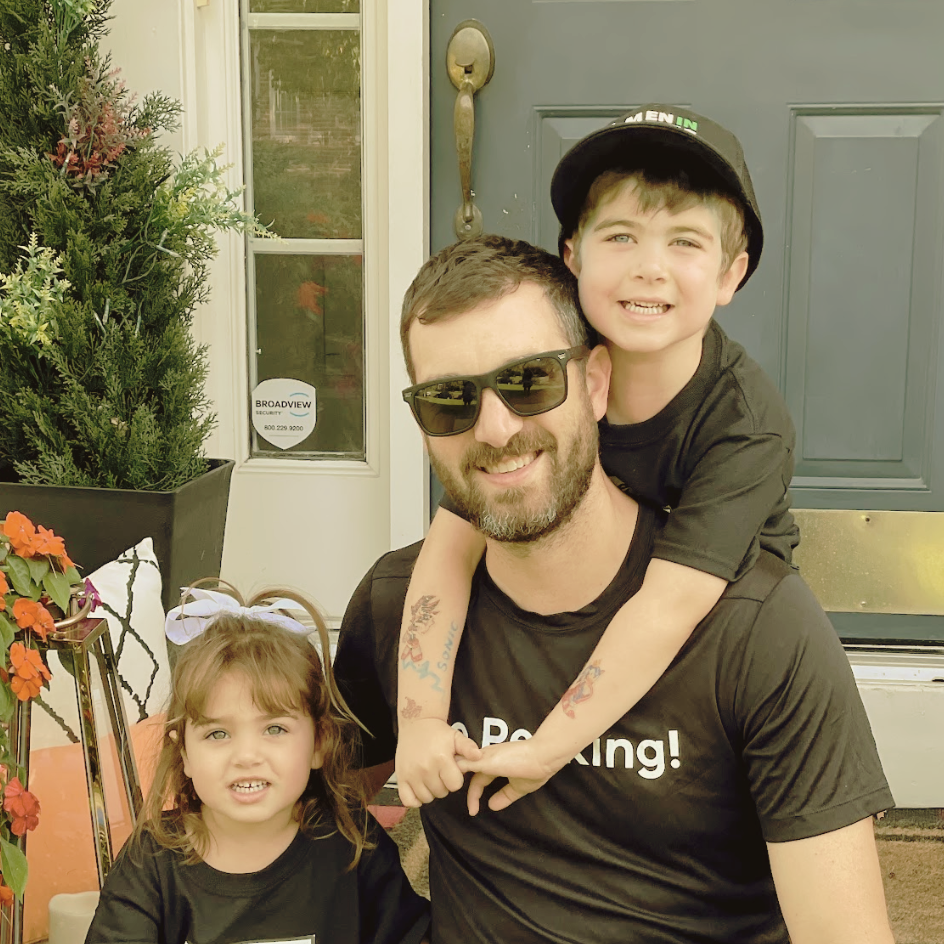 A man with sunglasses and a beard, a young boy on his shoulders, and a young girl standing next to him outside a house near the door with a black door and window. The girl has a white bow in her hair, and the boy is smiling and wearing a black cap. A potted plant with orange flowers is visible on the left.
