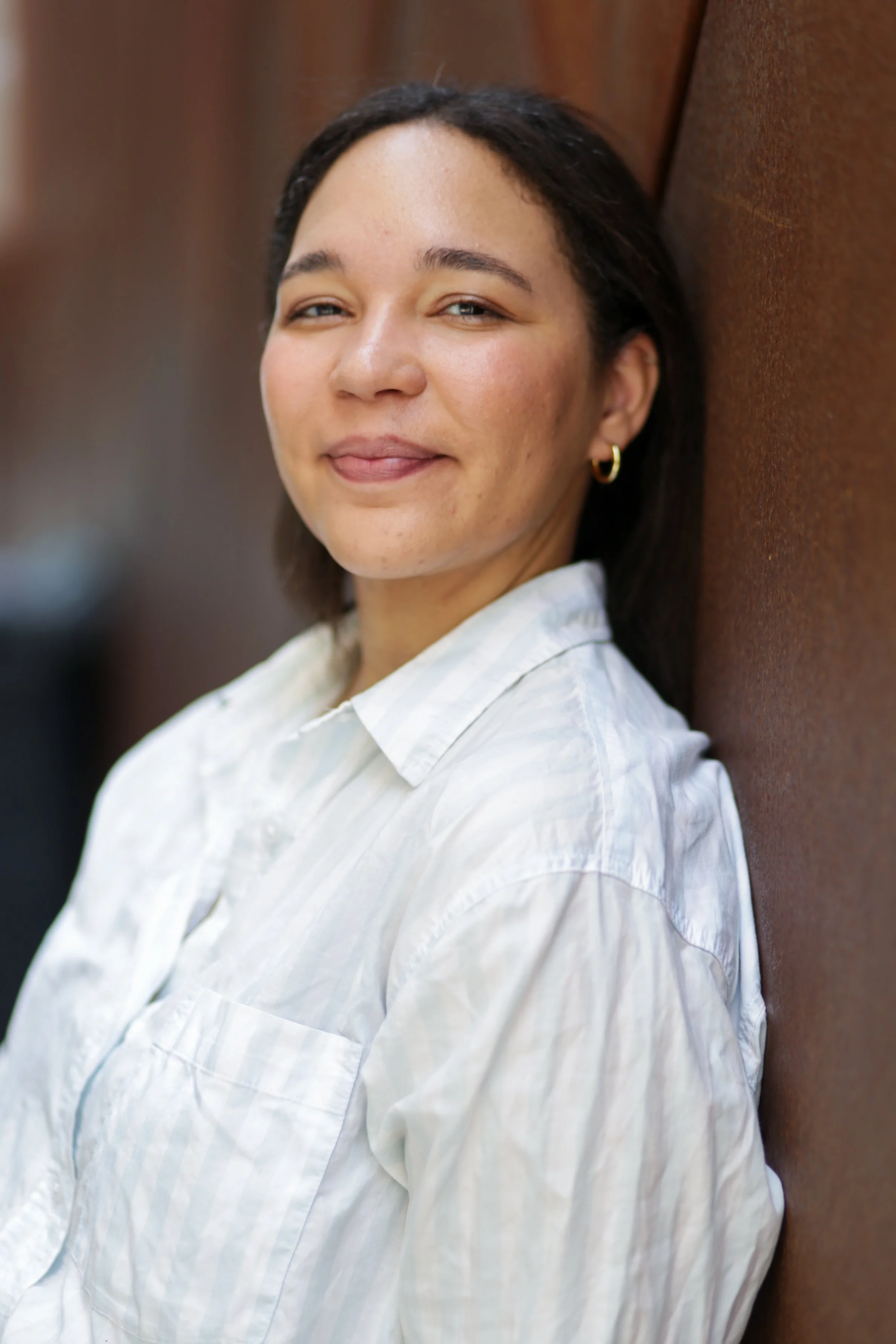 A young woman with dark hair, wearing a white button-up shirt and small hoop earrings, leaning against a brown textured wall, smiling at the camera.