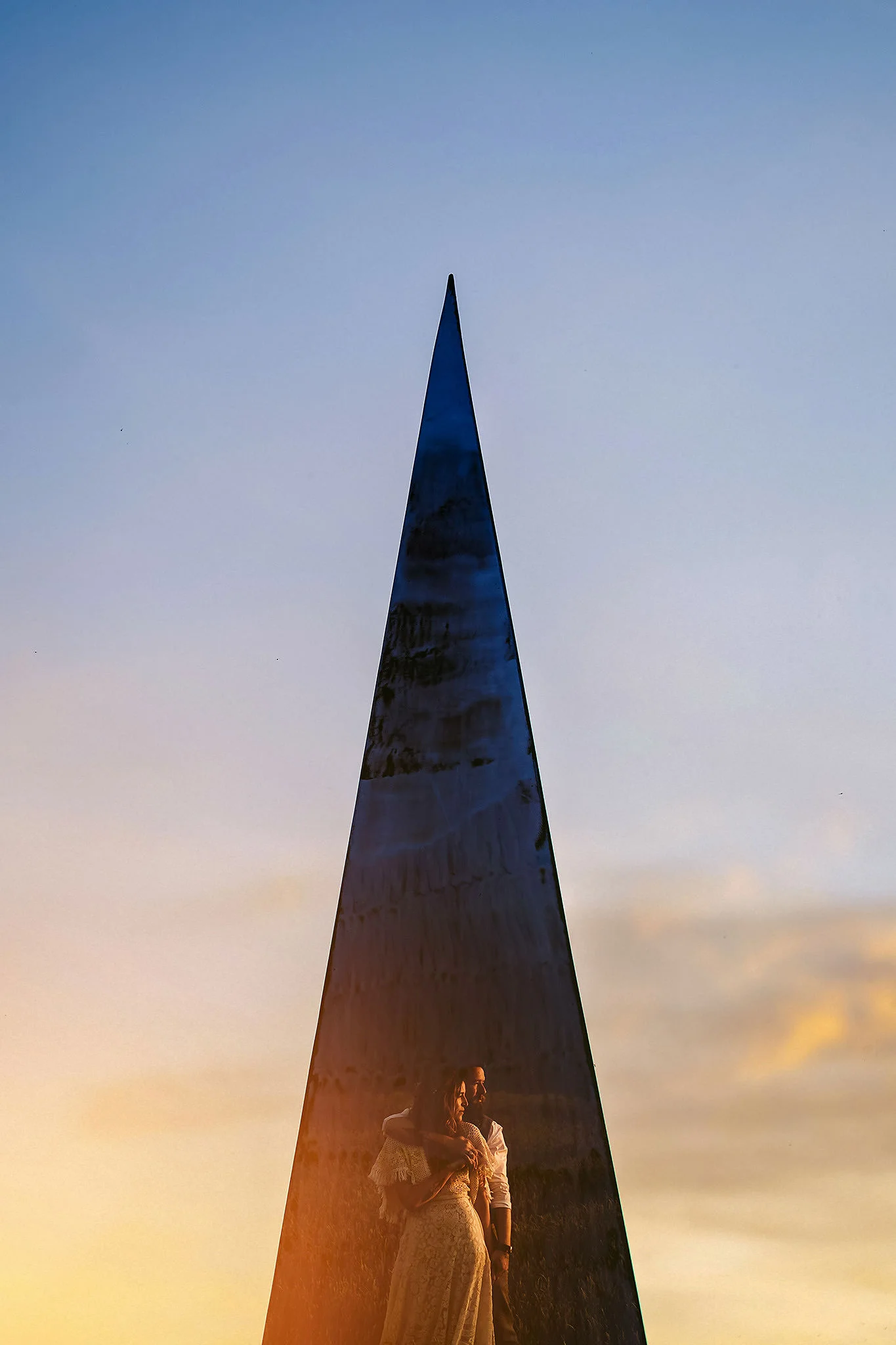 A bride and groom, standing close together in front of a tall, pointed metal sculpture at Field of Dreams in Northern Ireland during sunset with a cloudy sky in the background.