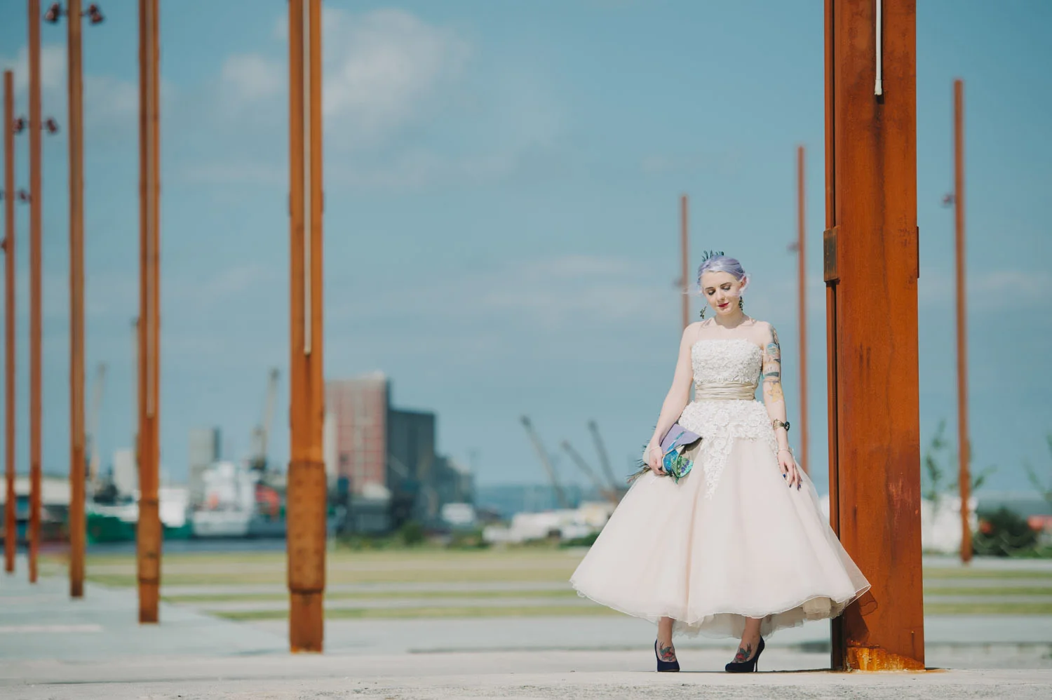 A bride with purple hair in a white wedding dress standing outdoors near large rust-colored metal beams on Titanic Slipway in Belfast