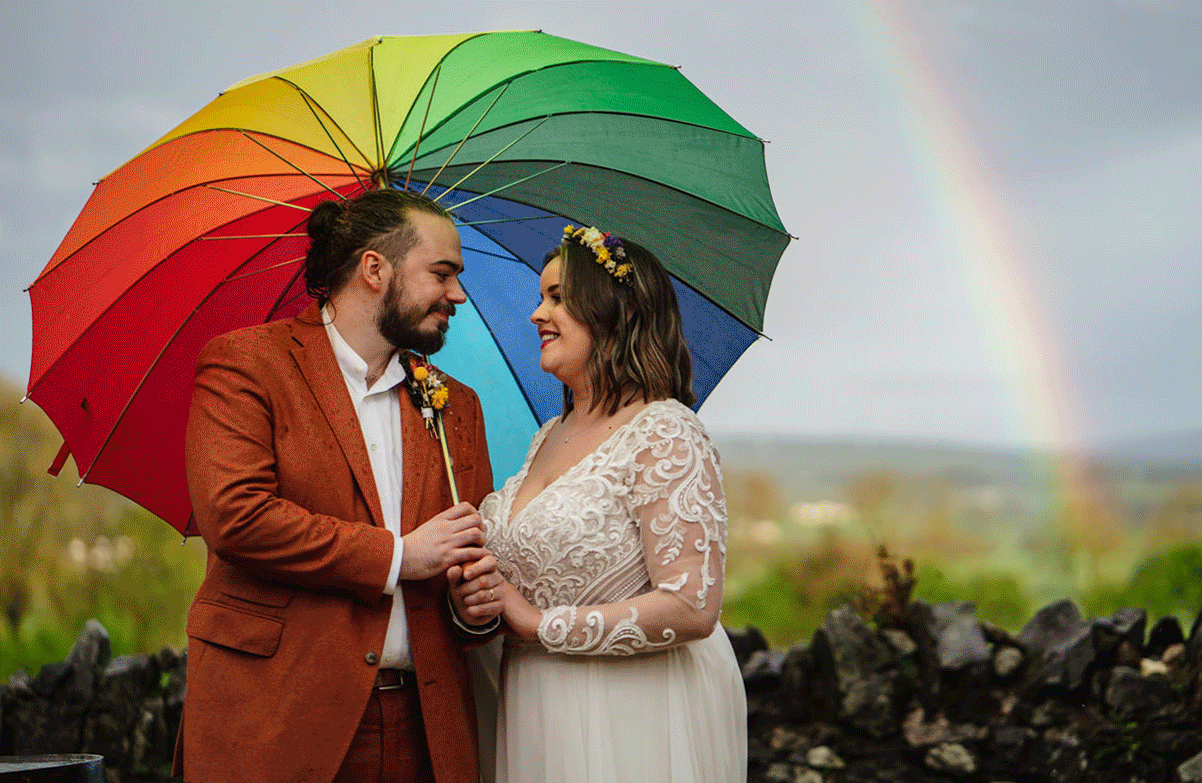 A couple posing with a multi-coloured umbrella with a rainbow in the background at Limepark in Northern Ireland