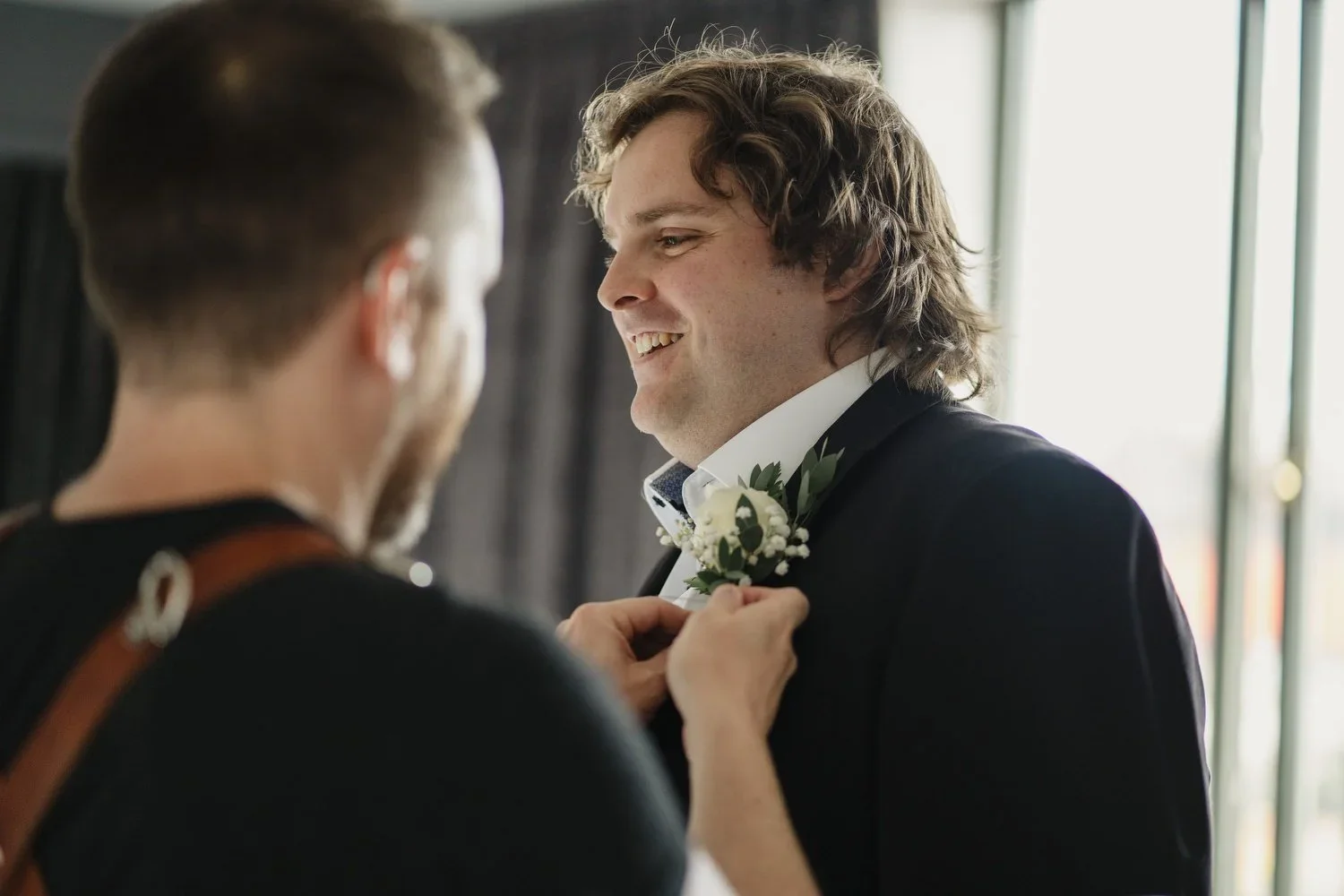 A man in a black suit with a white shirt smiling as another person pins a white flower boutonniere on his lapel, near a window with light streaming in.