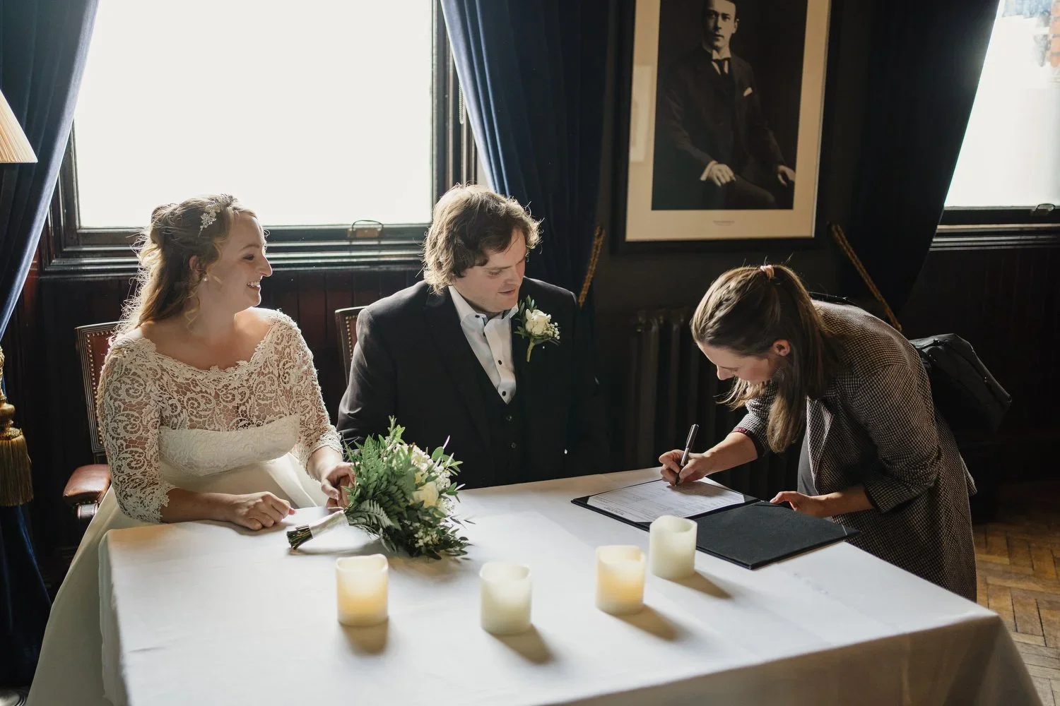 A bride and groom sit at a wedding ceremony table with a woman signing a document. The bride is smiling and has a bouquet of flowers, the groom is looking at the document, and the woman is focused on signing. Candles are on the table, and a framed portrait hangs on the wall behind them in a room with dark wood paneling and blue curtains.
