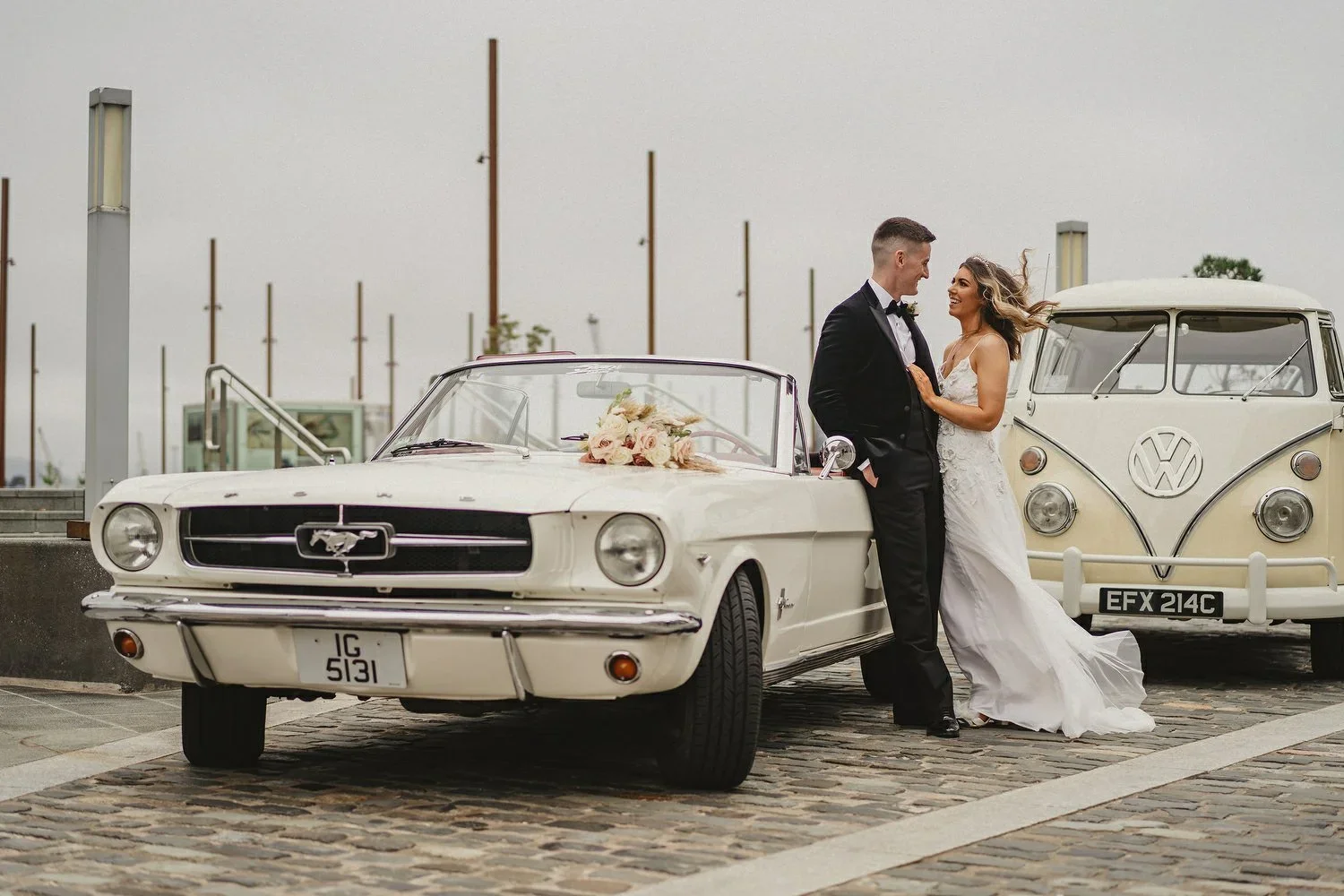 A bride and groom pose with a vintage Ford Mustang and VW Camper outside the Titanic hotel in Belfast on a rainy wedding day