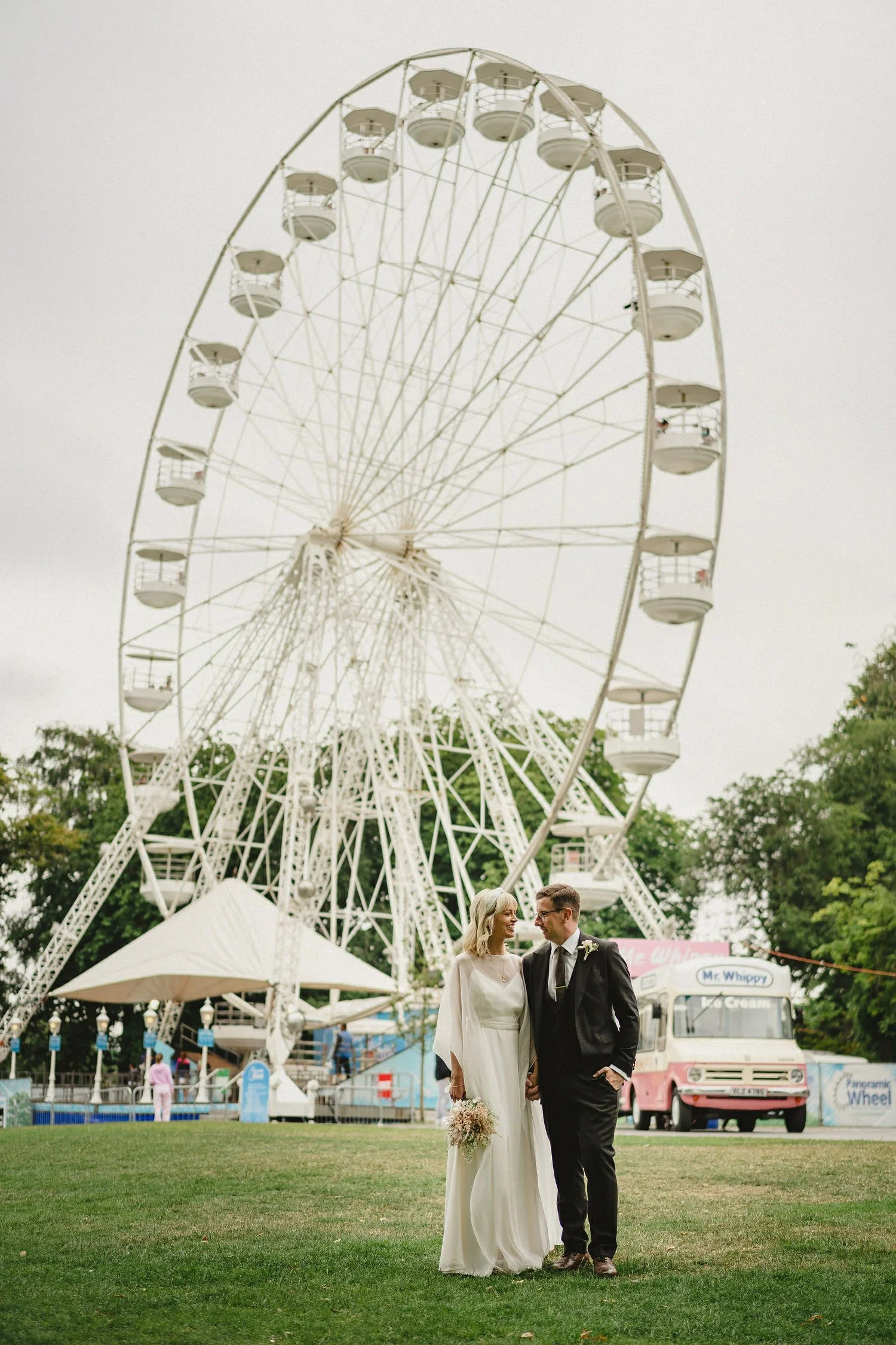 A bride and groom pose in front of a ferris wheel in Belfast Botanic Gardens
