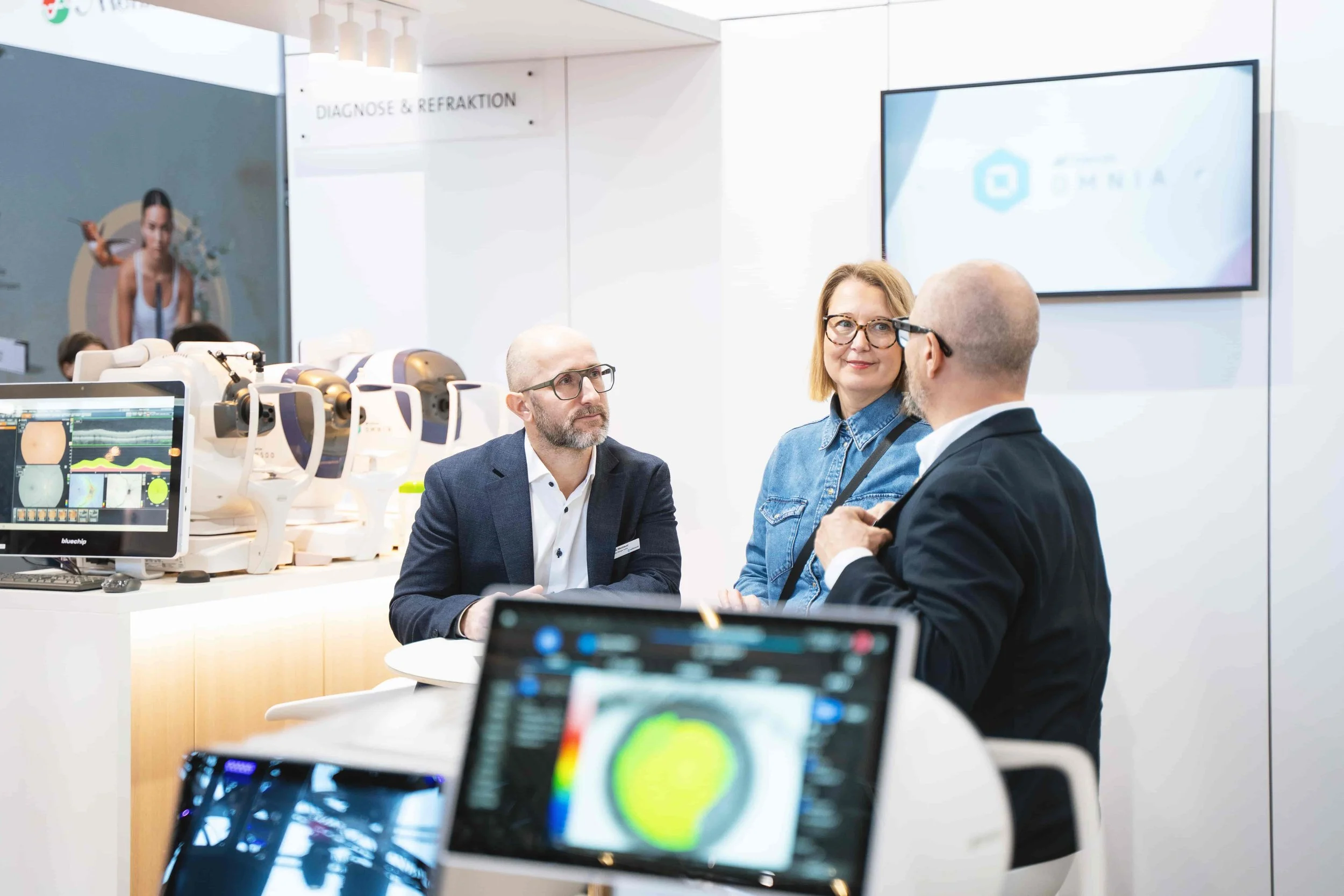 Three people engaged in a discussion at a technology exhibition. In the background, there are medical or scientific devices and a large screen displaying a logo. The setting appears to be an exhibition or conference booth for medical or technological innovation.