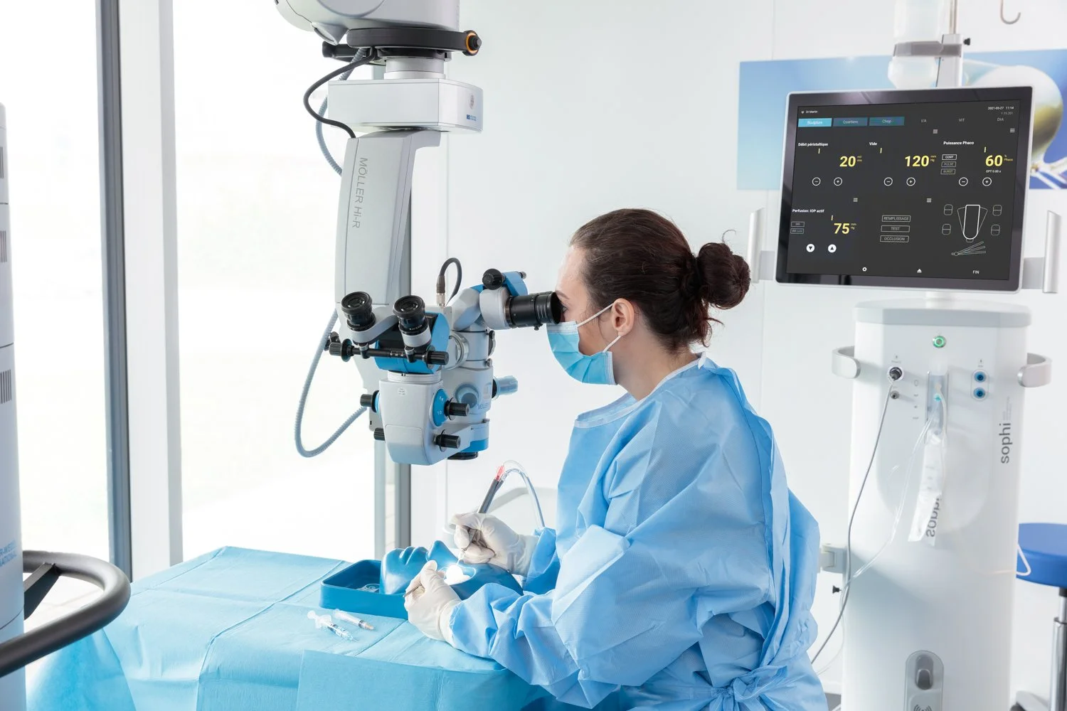 A medical professional wearing a face mask and protective gown, using a microscope and a surgical instrument in a clinical setting with advanced medical equipment.