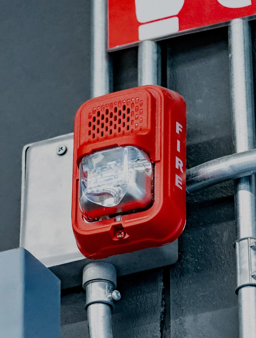 Red fire alarm with a clear plastic cover mounted on a gray wall near metallic conduit pipes.