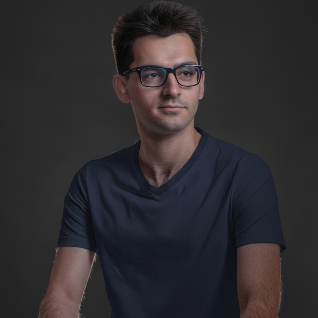 A young man with dark hair, wearing glasses and a navy blue T-shirt, looks off to the side against a dark gray background.