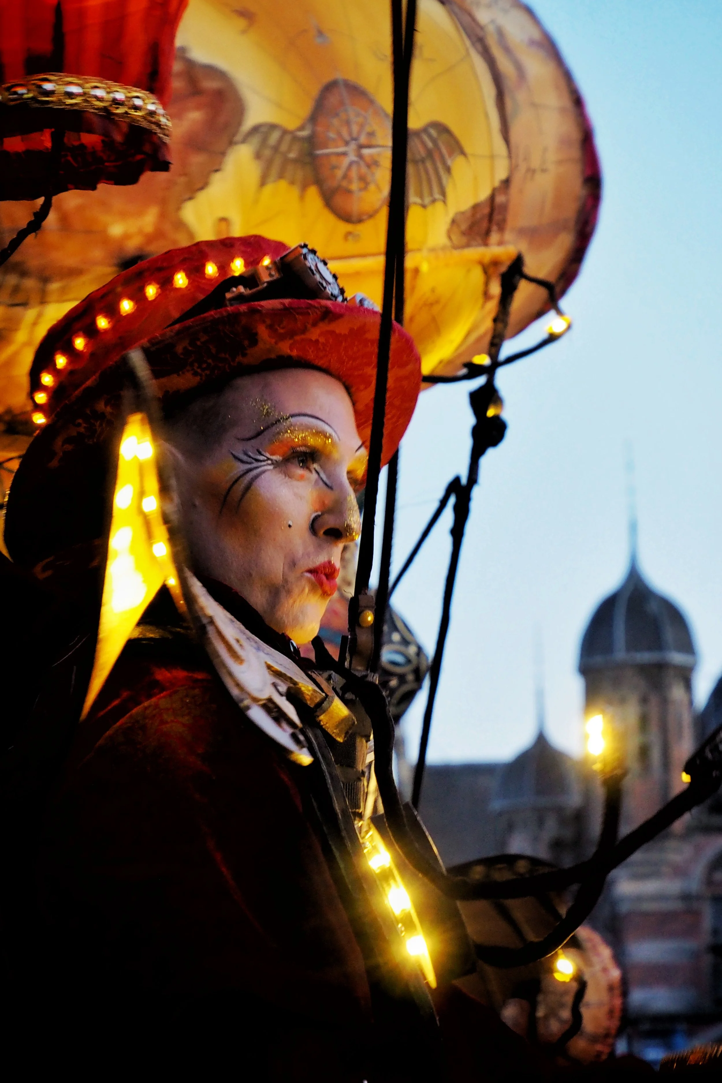 Close-up of a performer in colorful costume and makeup, holding a decorated umbrella with yellow lights, against a backdrop of historic buildings topped with domes at dusk.